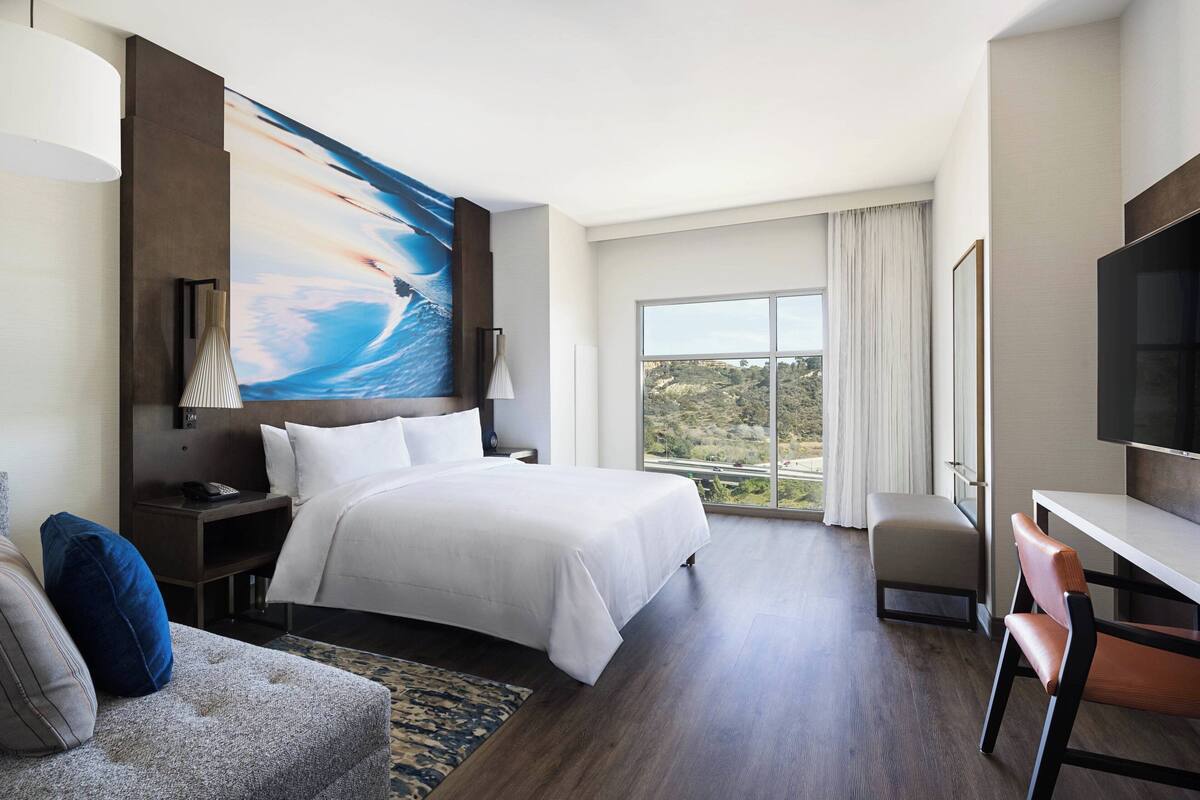 A modern hotel room with a large bed, a desk, a TV, and a view of the outdoors.