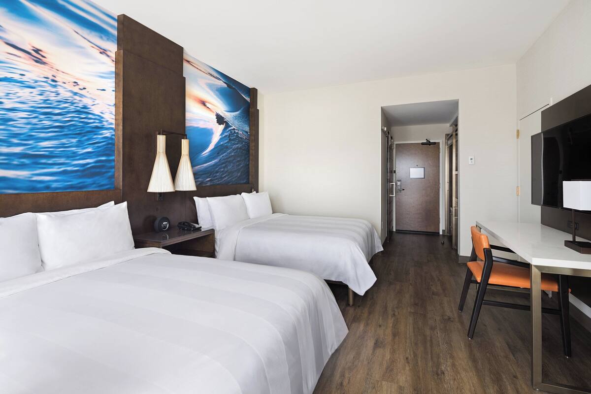 A hotel room with two beds, a desk, a chair, a TV, and a painting of a seascape.