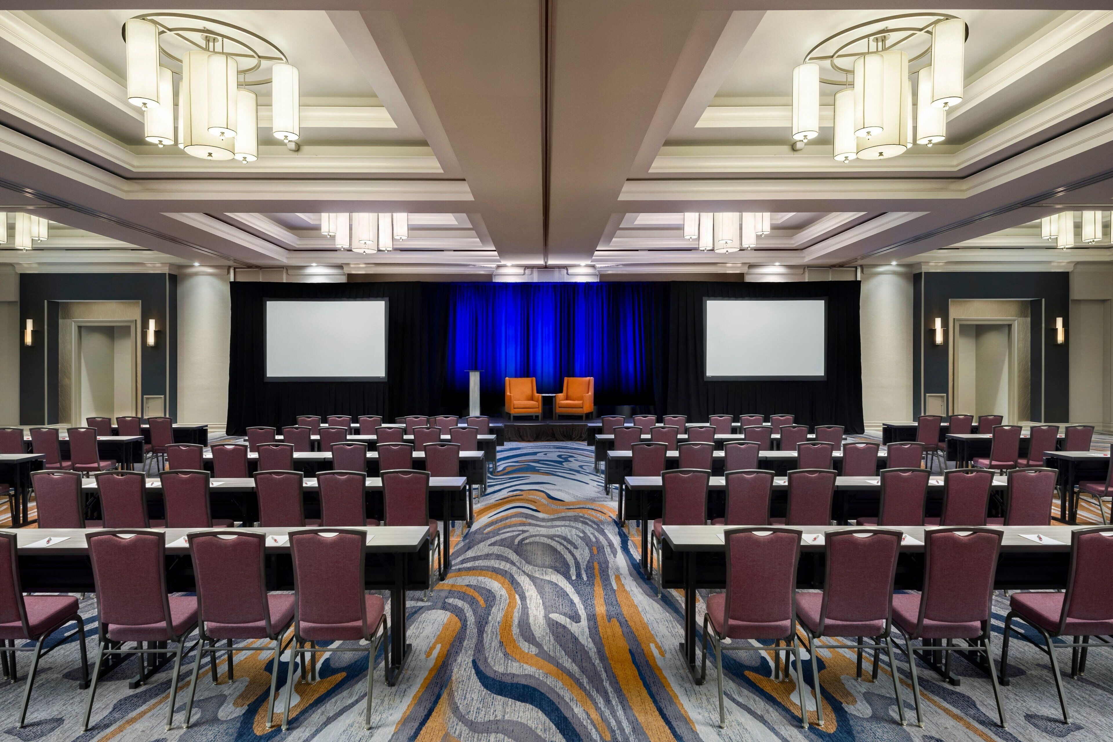 A conference room with rows of chairs, a stage with a blue curtain, and a large screen.