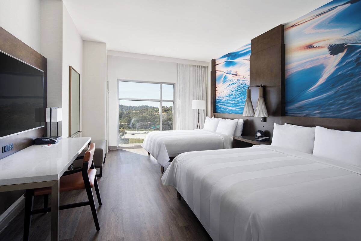 A hotel room with two beds, a desk, a TV, and a large window with a view of the outdoors.