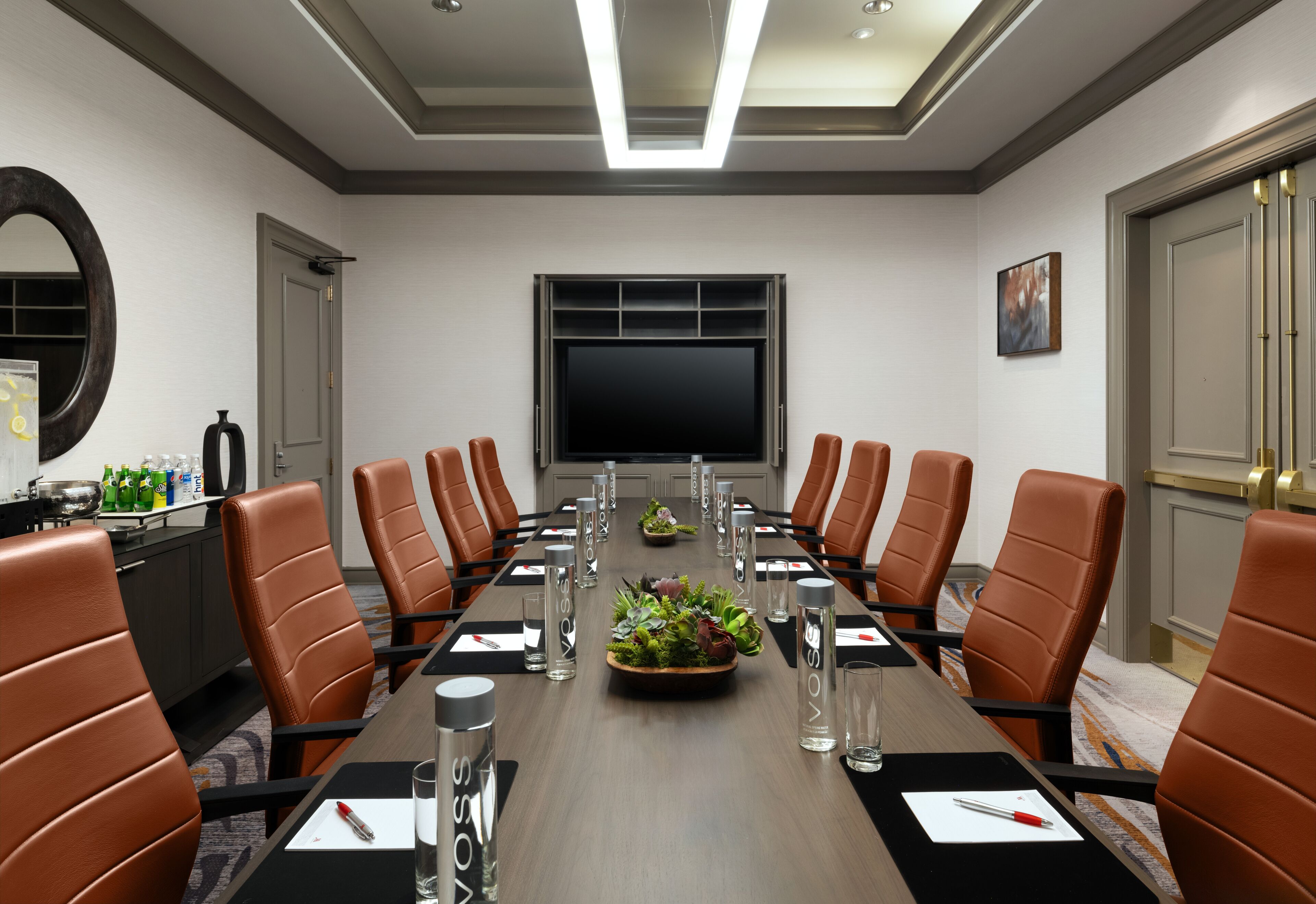 A conference room with a long table, brown leather chairs, a large screen, and a plant centerpiece.