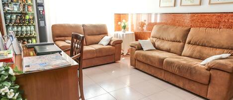 A spacious living area with a brown sofa set, a wooden desk with a laptop and papers, a vending machine, and framed artwork on the wall.