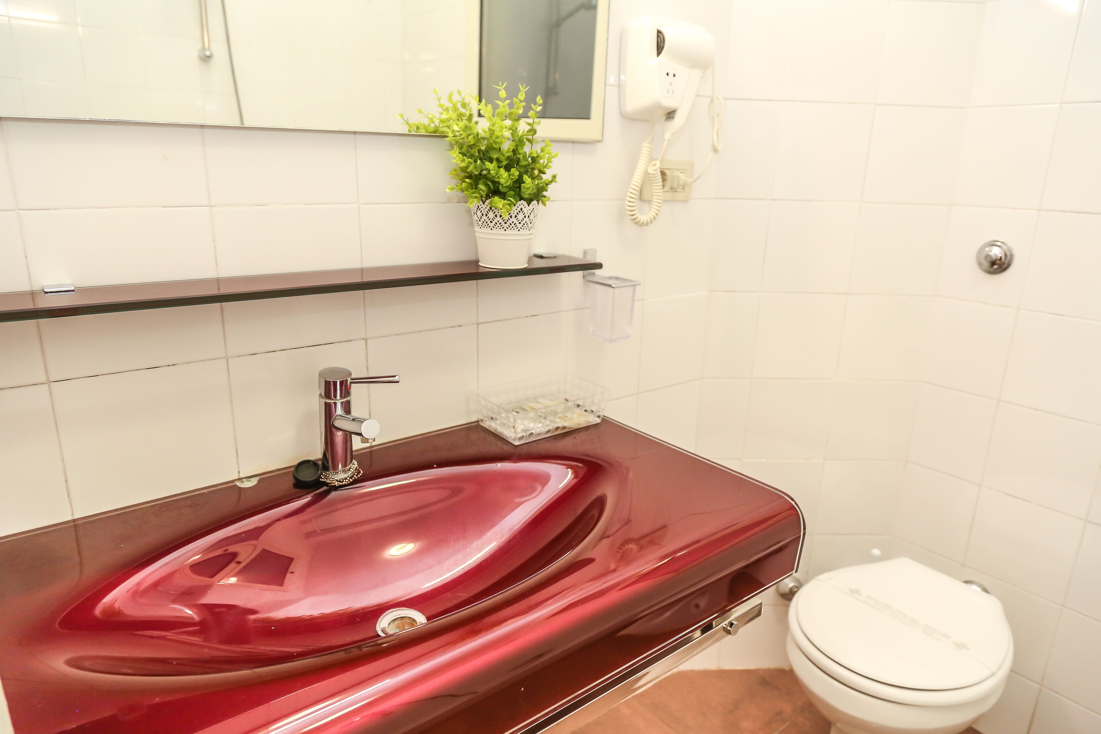 A bathroom with a red sink, white tiles, a small potted plant, a hairdryer, and a toilet.