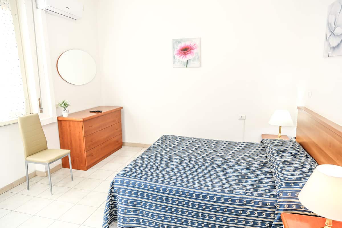 A bedroom with a bed, a wooden dresser, a chair, a round mirror, and a wall-mounted lamp.