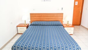 Un dormitorio con cabecera de madera, colcha estampada azul, dos mesitas de noche con lámparas, una pared blanca con una obra de arte floral y una puerta de madera con un cartel que dice 'F'.
