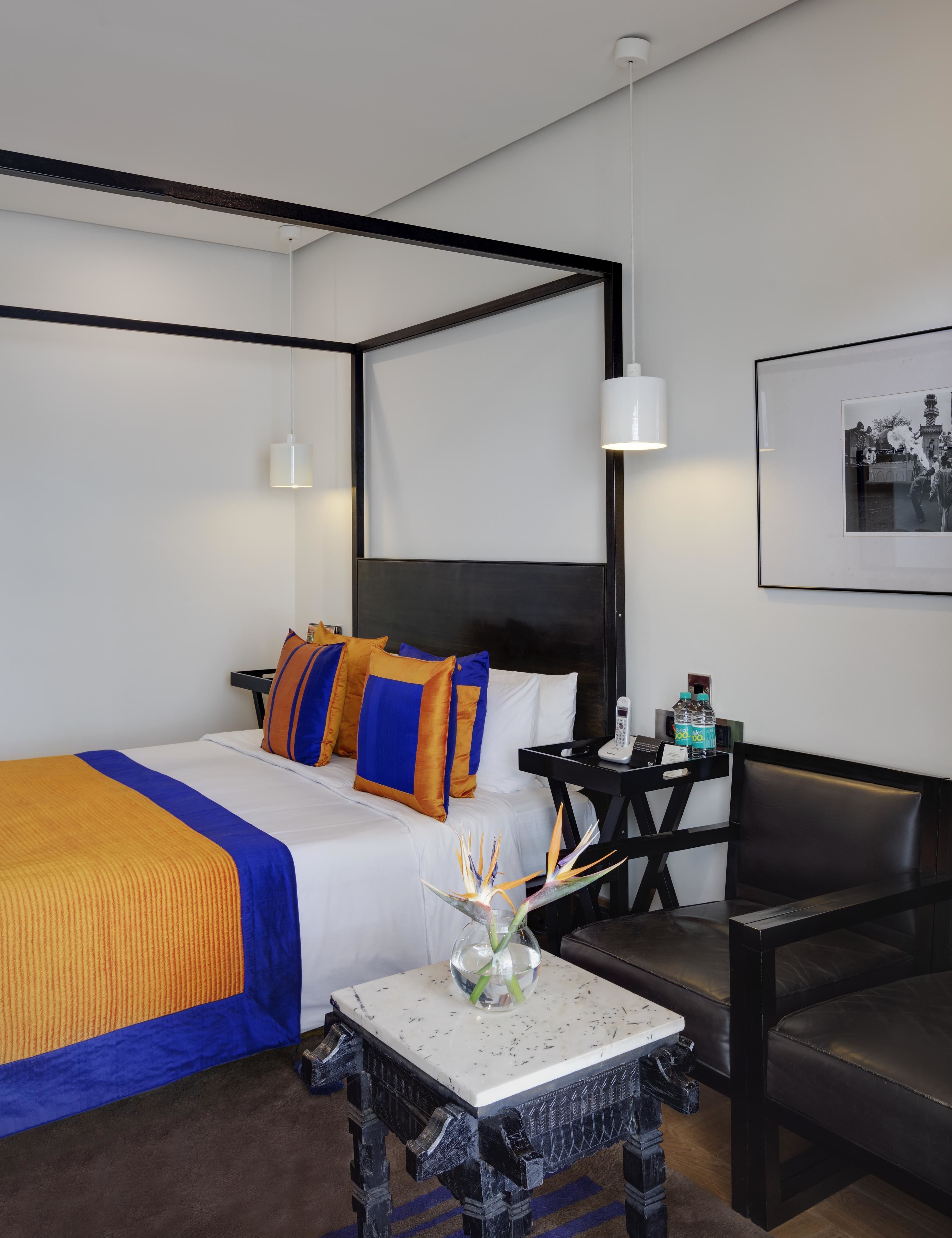 Una habitación de hotel moderna con una cama con dosel, una mesita con un florero, una silla y una mesita de noche con un teléfono y botellas de agua.