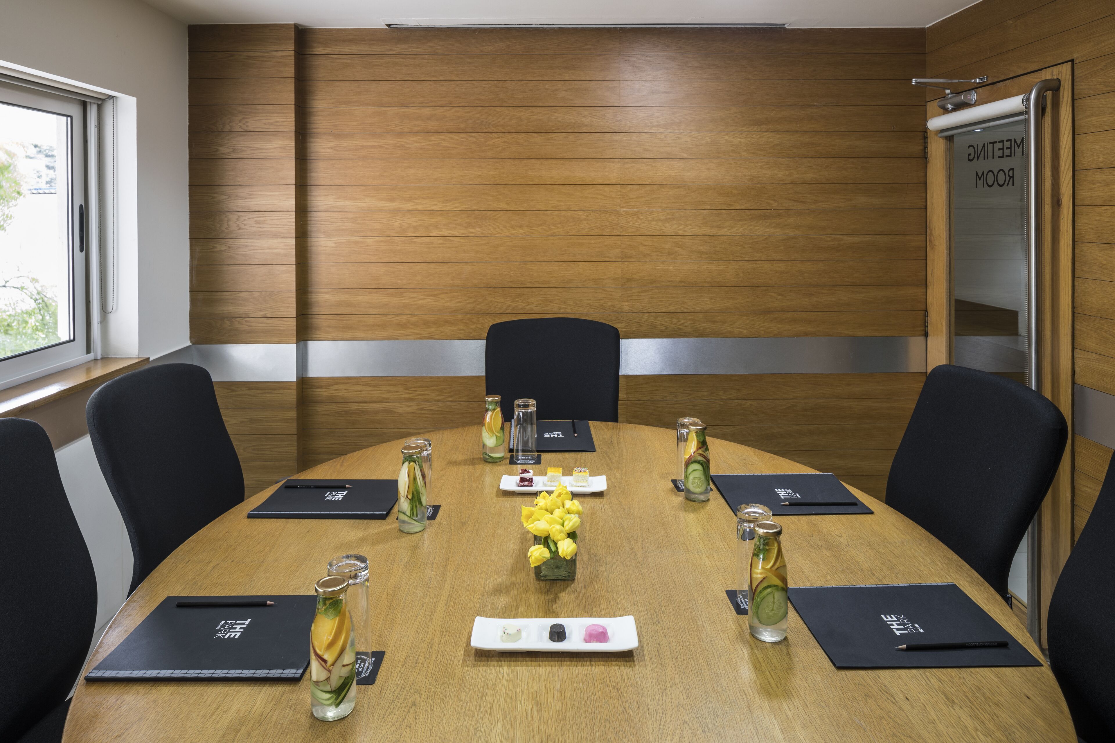Una sala de conferencias con pared de paneles de madera, sillas negras, una mesa redonda con tarjetas de identificación, un jarrón de flores amarillas y una botella de vidrio.