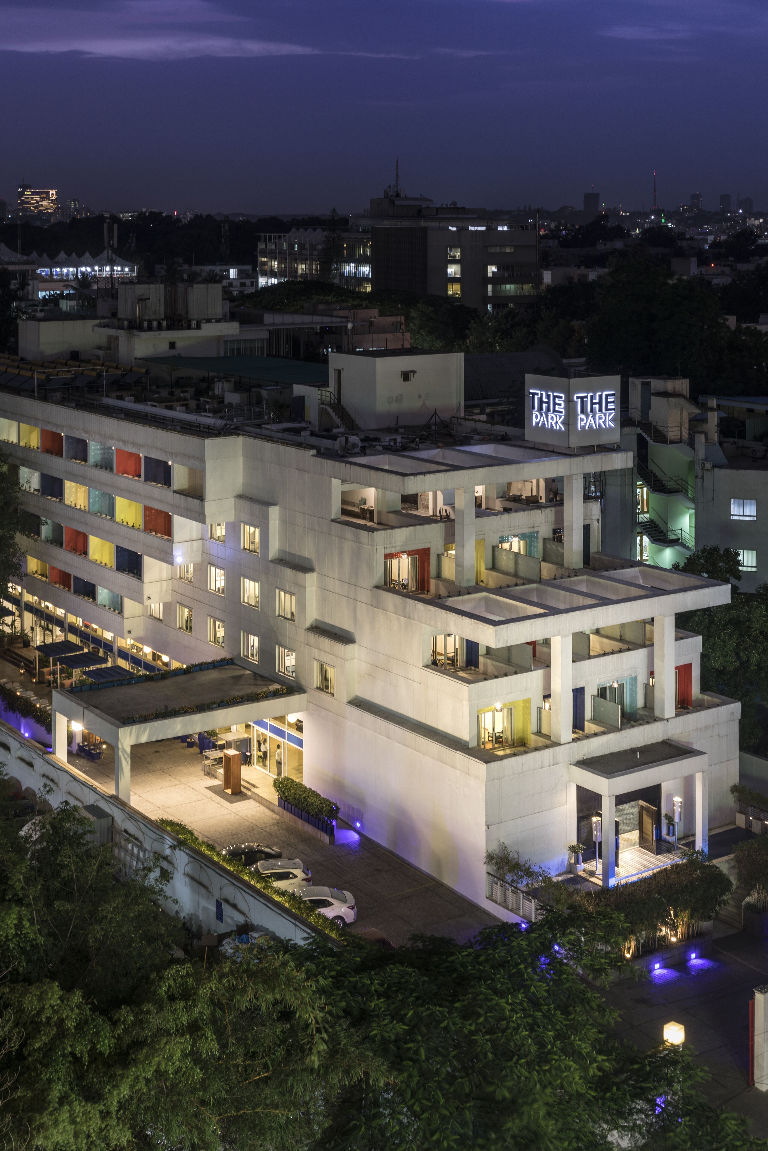 A multi-story hotel with a rooftop terrace, a sign reading "The Park," and a parking area with cars.