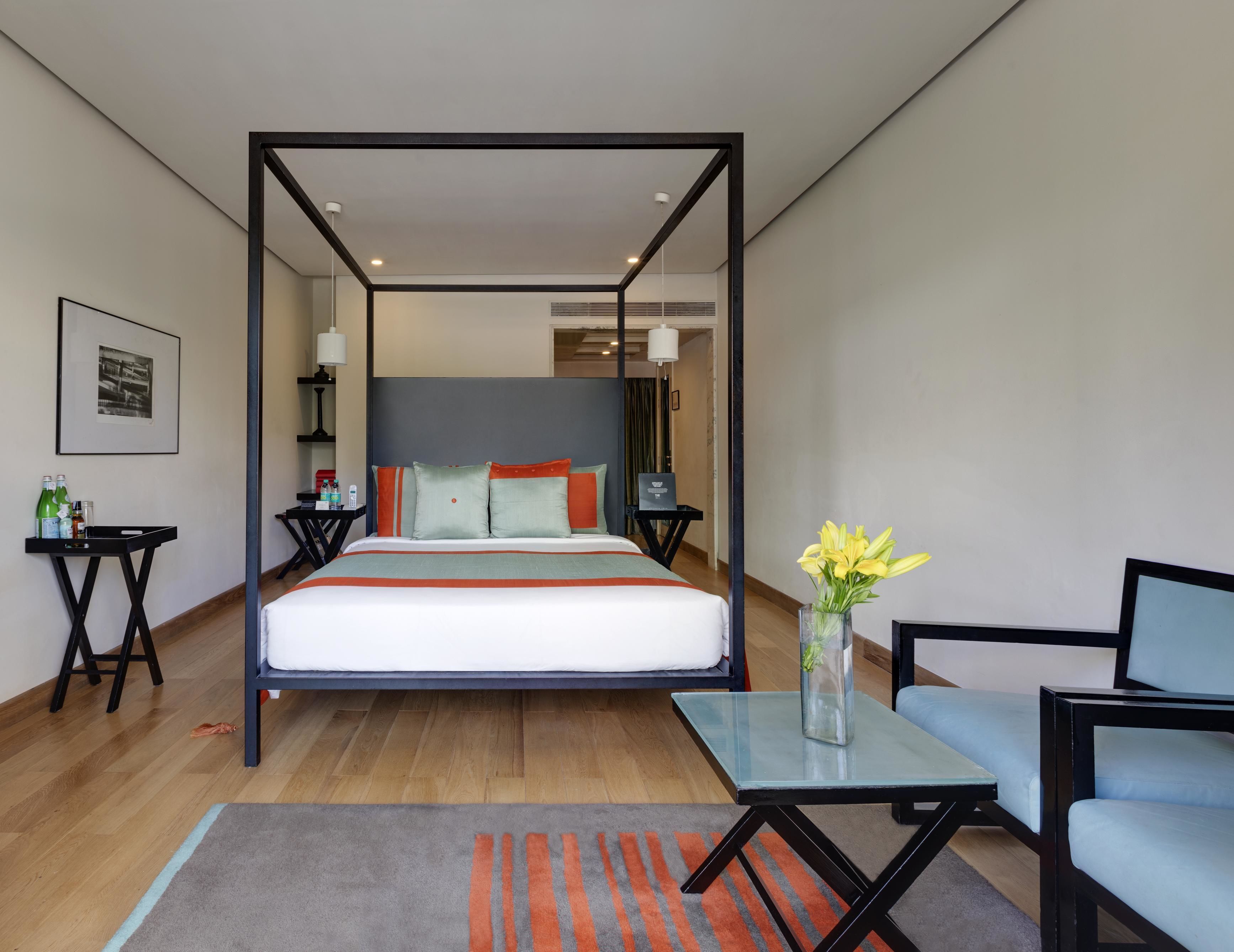 A modern hotel room with a four-poster bed, a glass coffee table, a blue armchair, and a small table with toiletries.