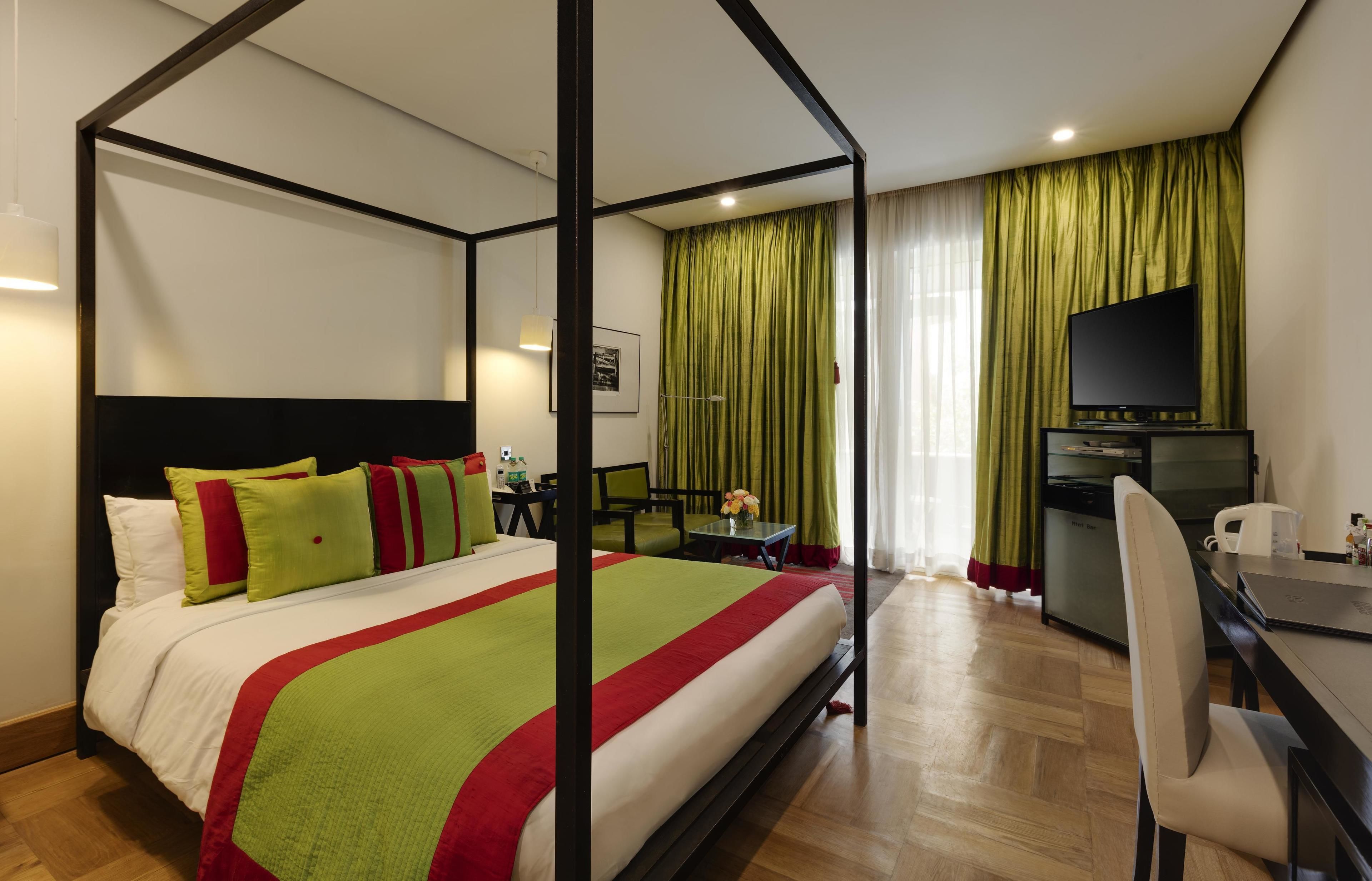 A four-poster bed with a red and green bedspread, a desk with a chair, a television, and a kitchen area with a refrigerator and microwave.