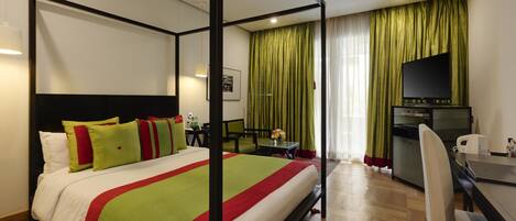 A four-poster bed with a red and green bedspread, a desk with a chair, a television, and a kitchen area with a refrigerator and microwave.