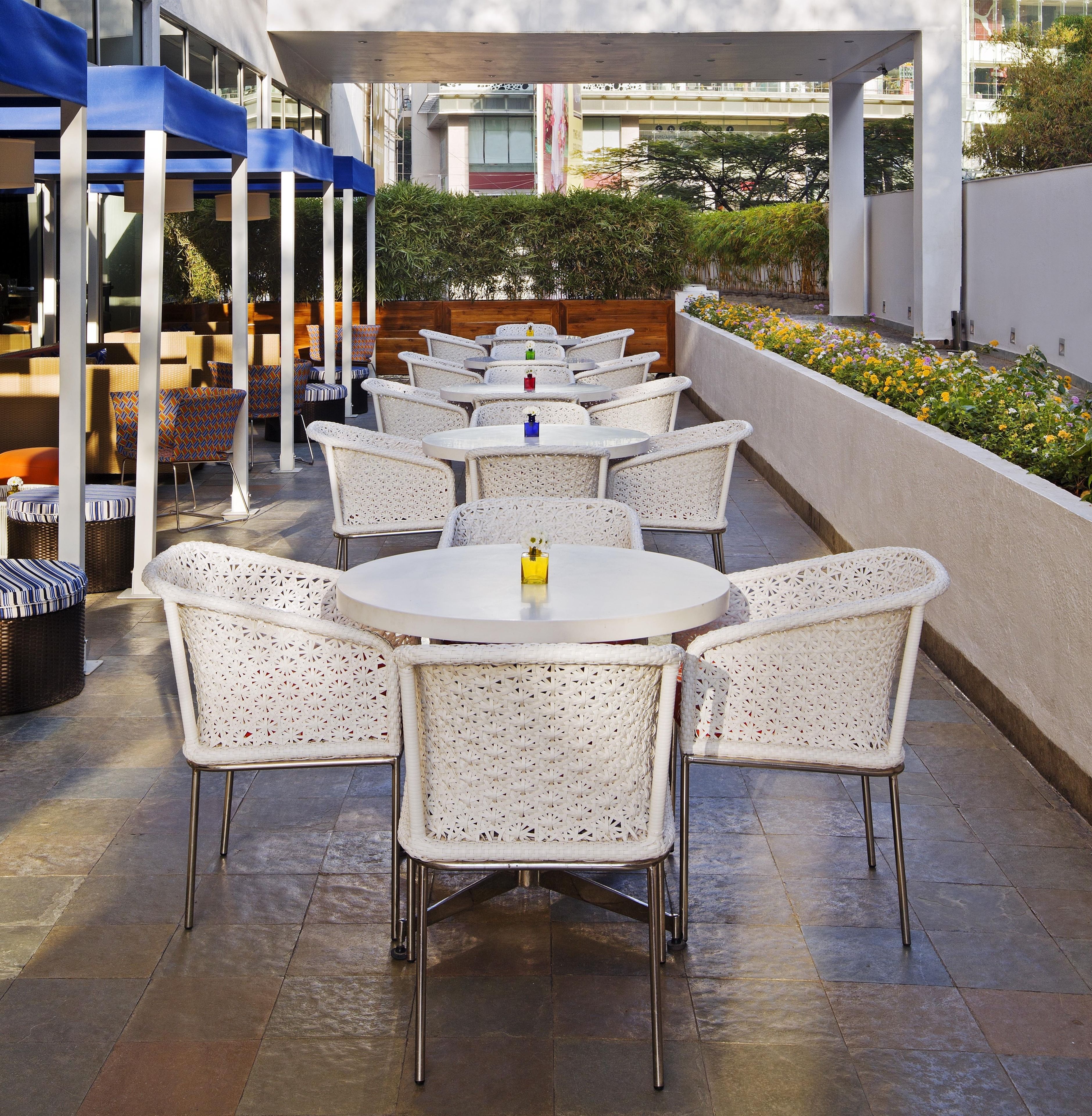 A covered outdoor seating area with white wicker chairs and round tables, featuring a view of a building and a garden in the background.