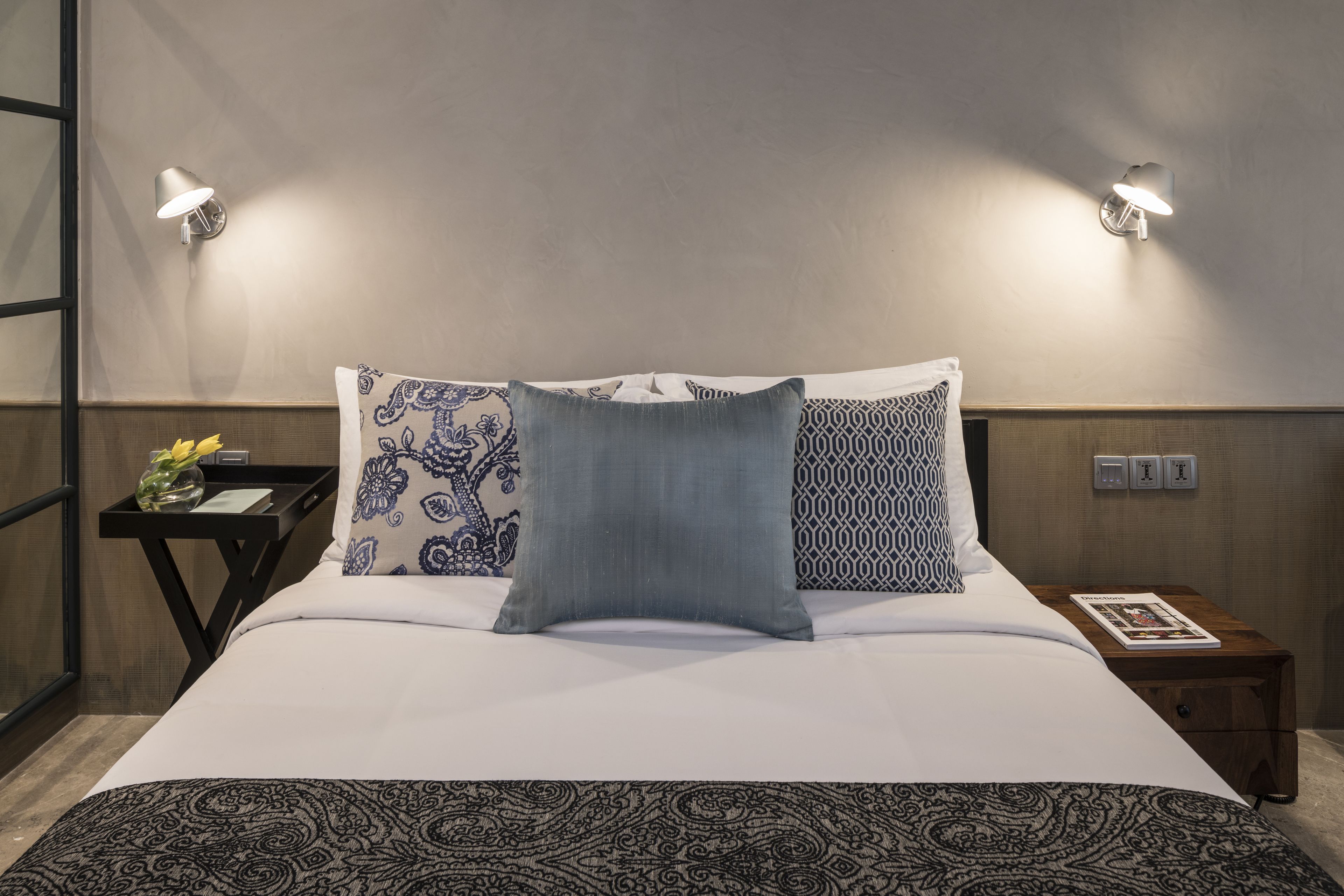 Una cama bien hecha con ropa de cama estampada, dos lámparas fijadas a la pared y una mesita de noche con una revista.