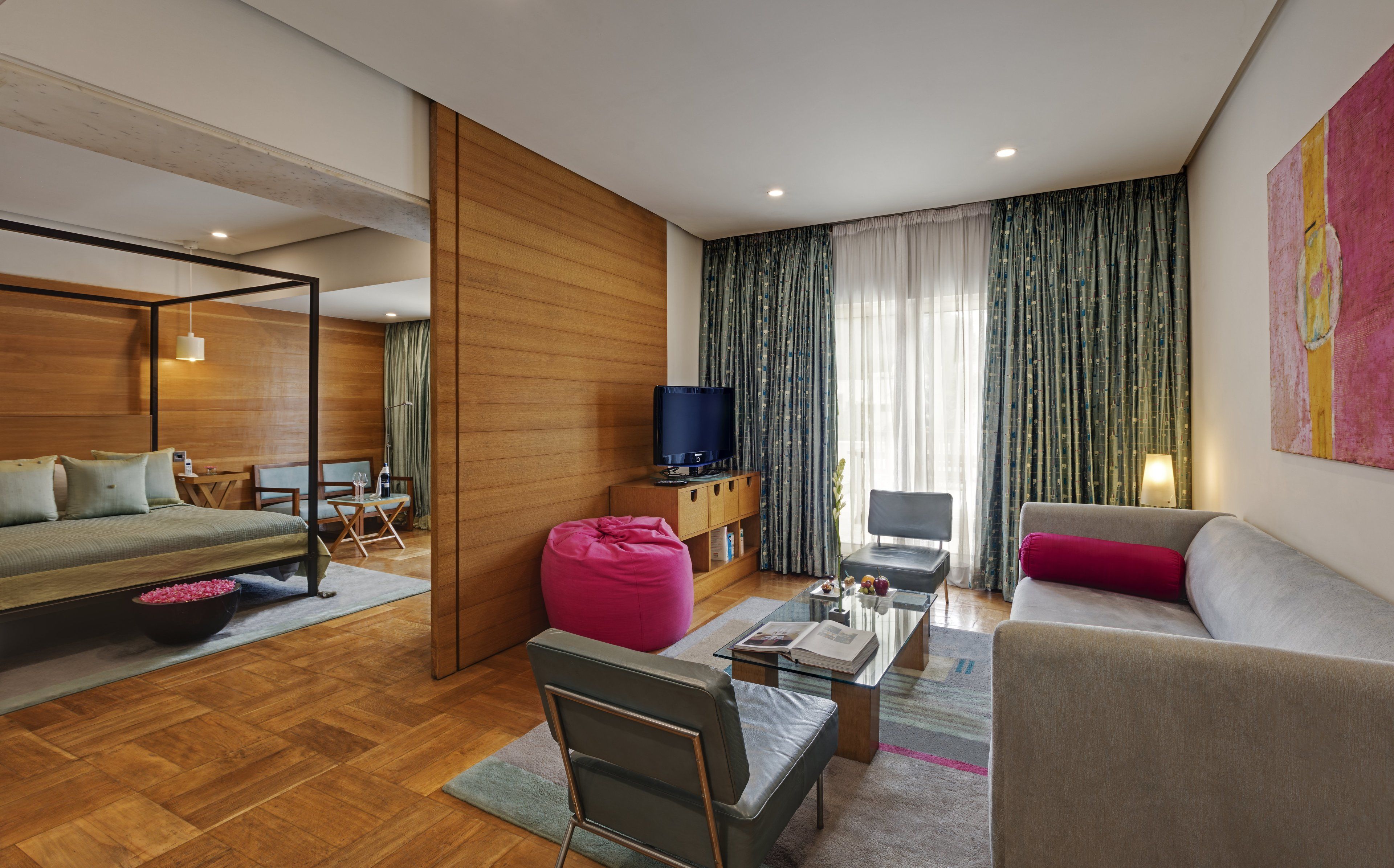 A modern hotel room with a bed, a sofa, a TV, a dining area, and a large window with curtains.