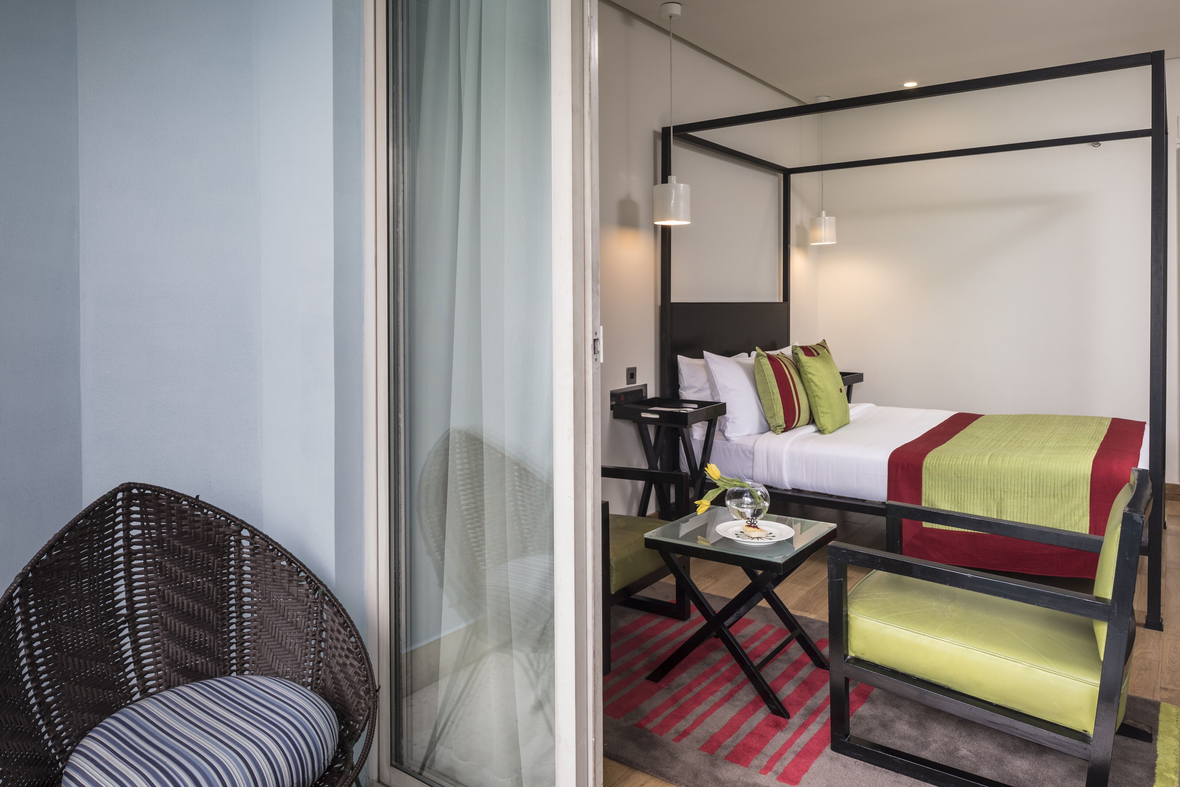 A hotel room with a four-poster bed, a wicker chair, a small table with a vase, and a red and gray striped rug.