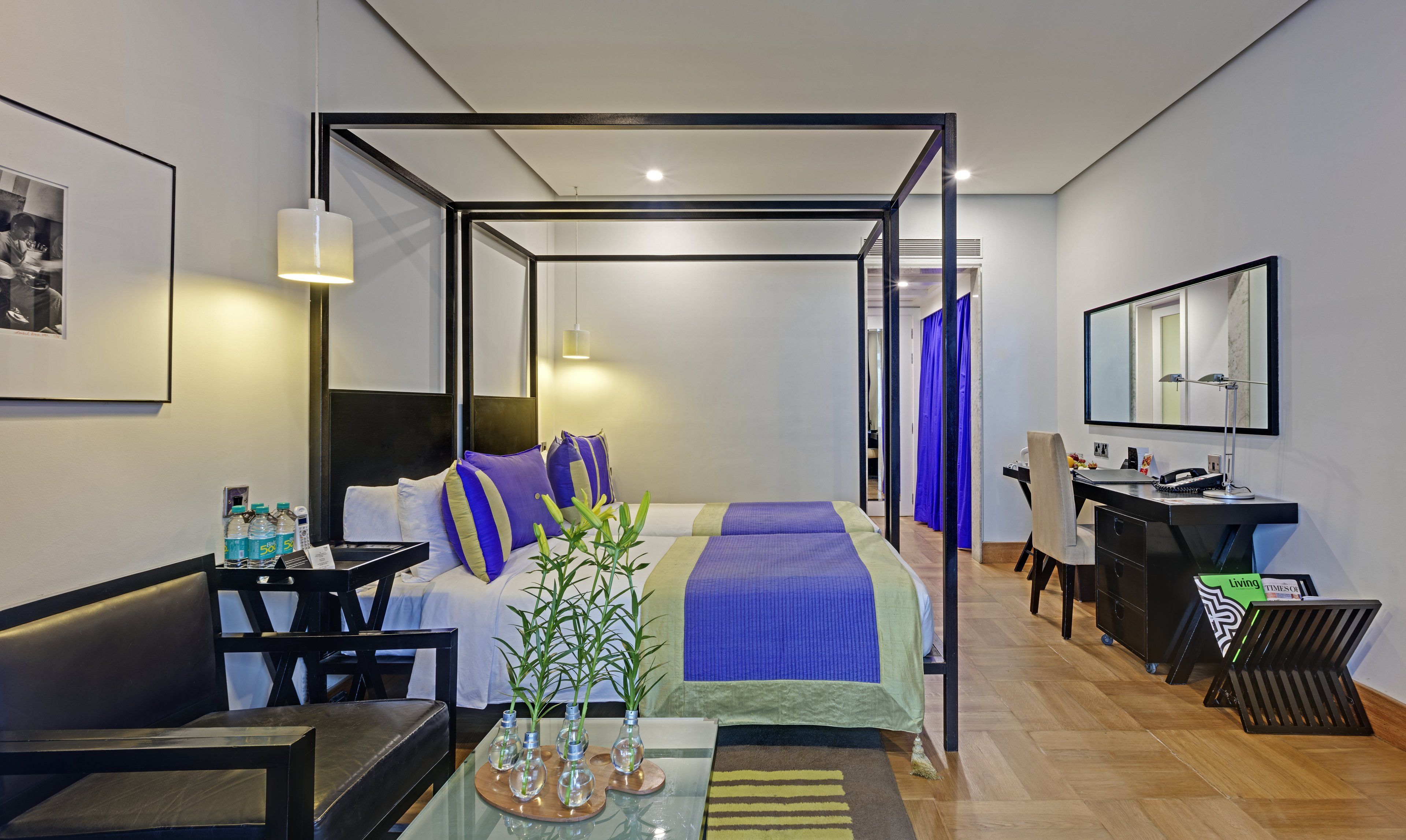 A modern hotel room with a bed, a desk, a chair, a mirror, and a bathroom visible through a glass partition.