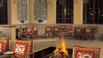 An outdoor dining area with wooden tables and chairs, a central fire pit, and decorative wall panels.