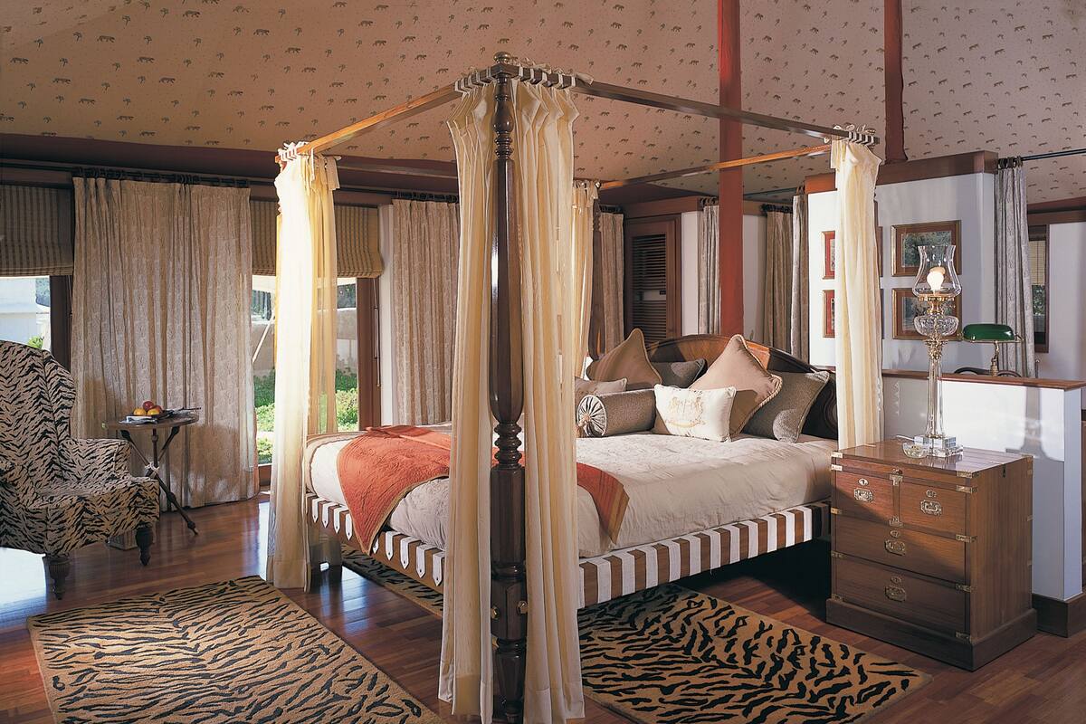 Un lit à baldaquin à quatre poteaux avec une tête de lit en bois, un tapis à motifs zébrés, une table de chevet en bois et une lampe verte.
