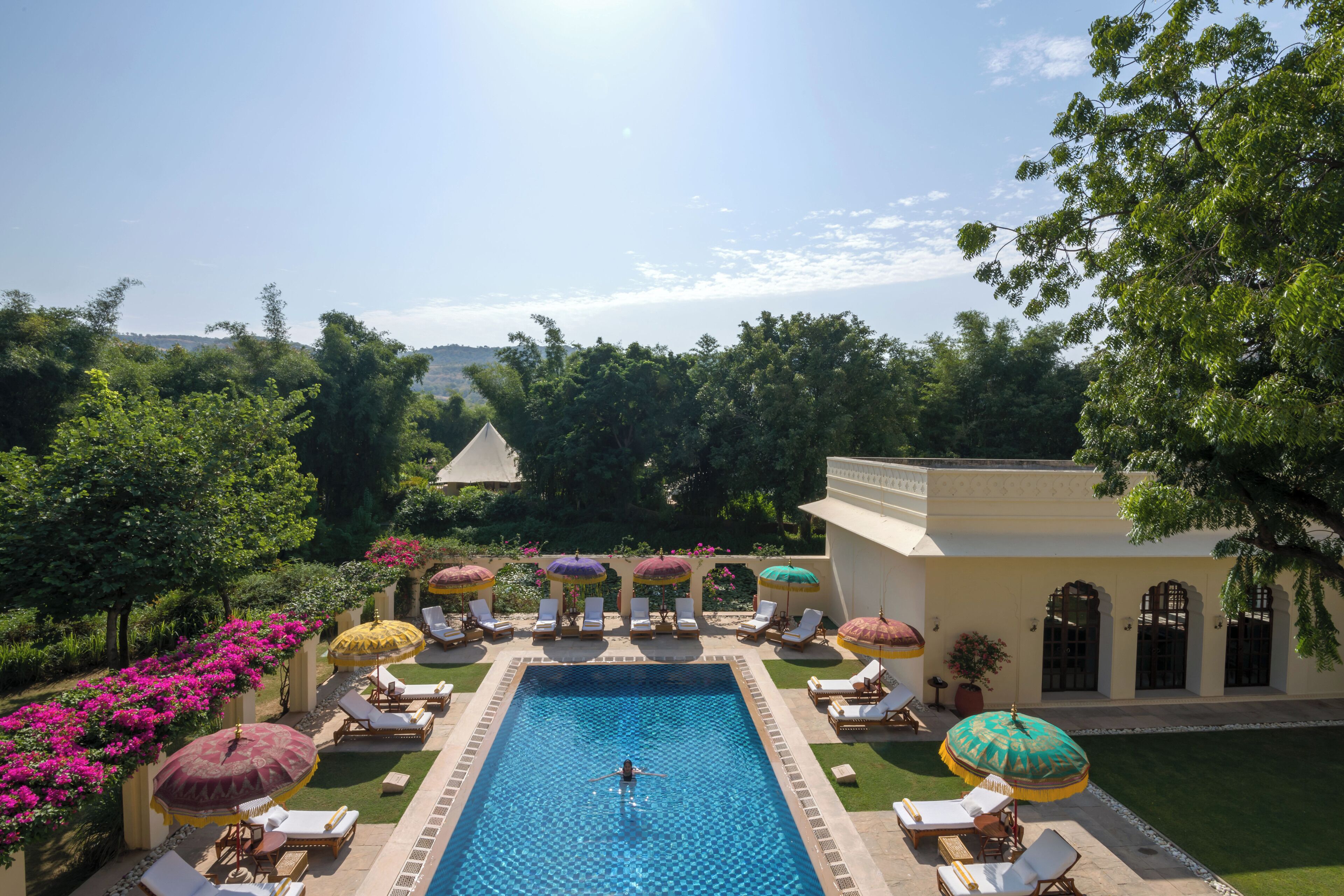 A pool area with lounge chairs and umbrellas, surrounded by greenery and a building with arches.