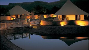 A serene evening scene at a resort with two large tents illuminated by warm lighting, surrounded by a reflective pool and a landscaped area with pathways and wooden fences. - The Oberoi Vanyavilas, Ranthambore (Sawai Madhopur)