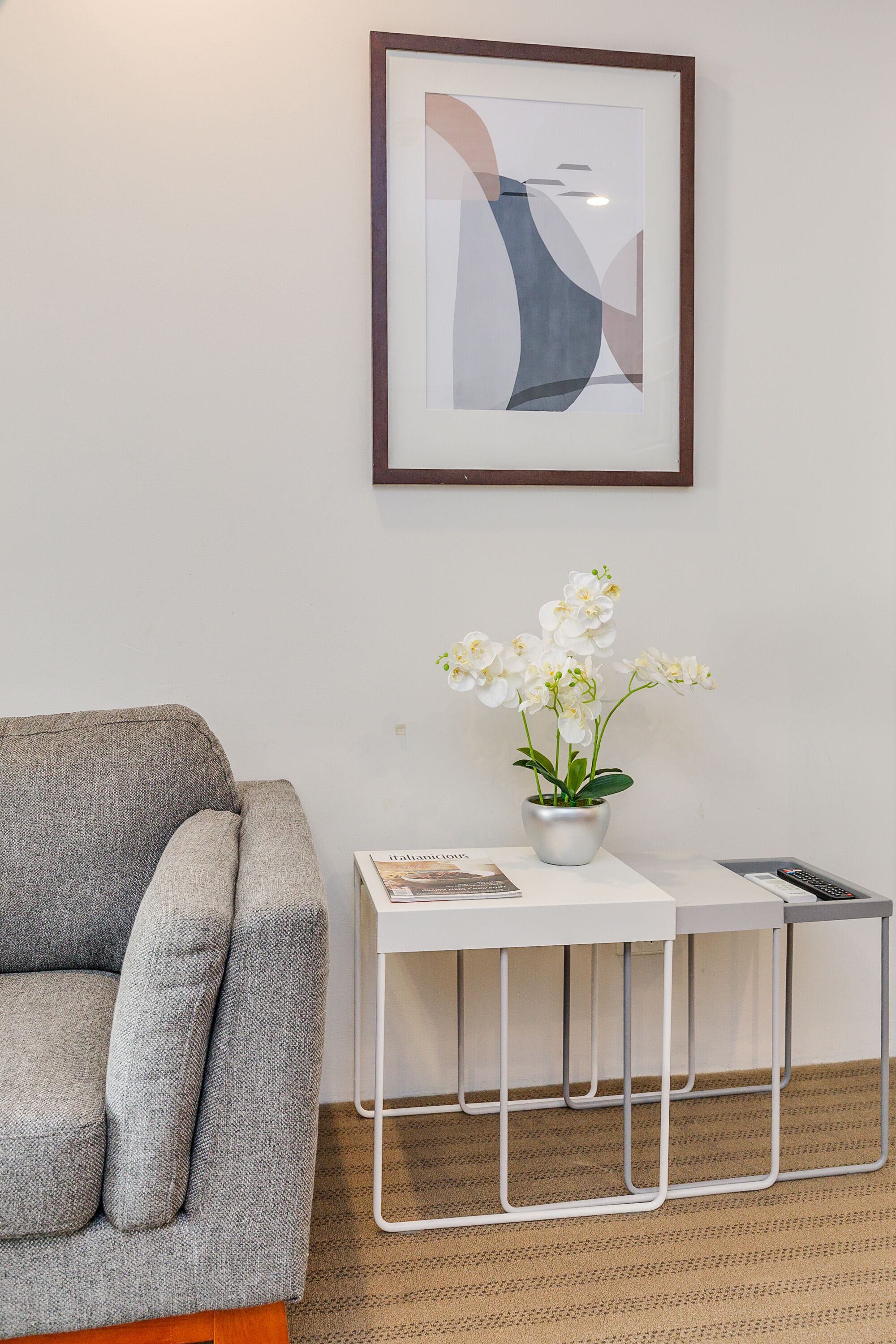 A modern living room with a grey sofa, a white side table with a potted orchid, and a framed abstract artwork on the wall.