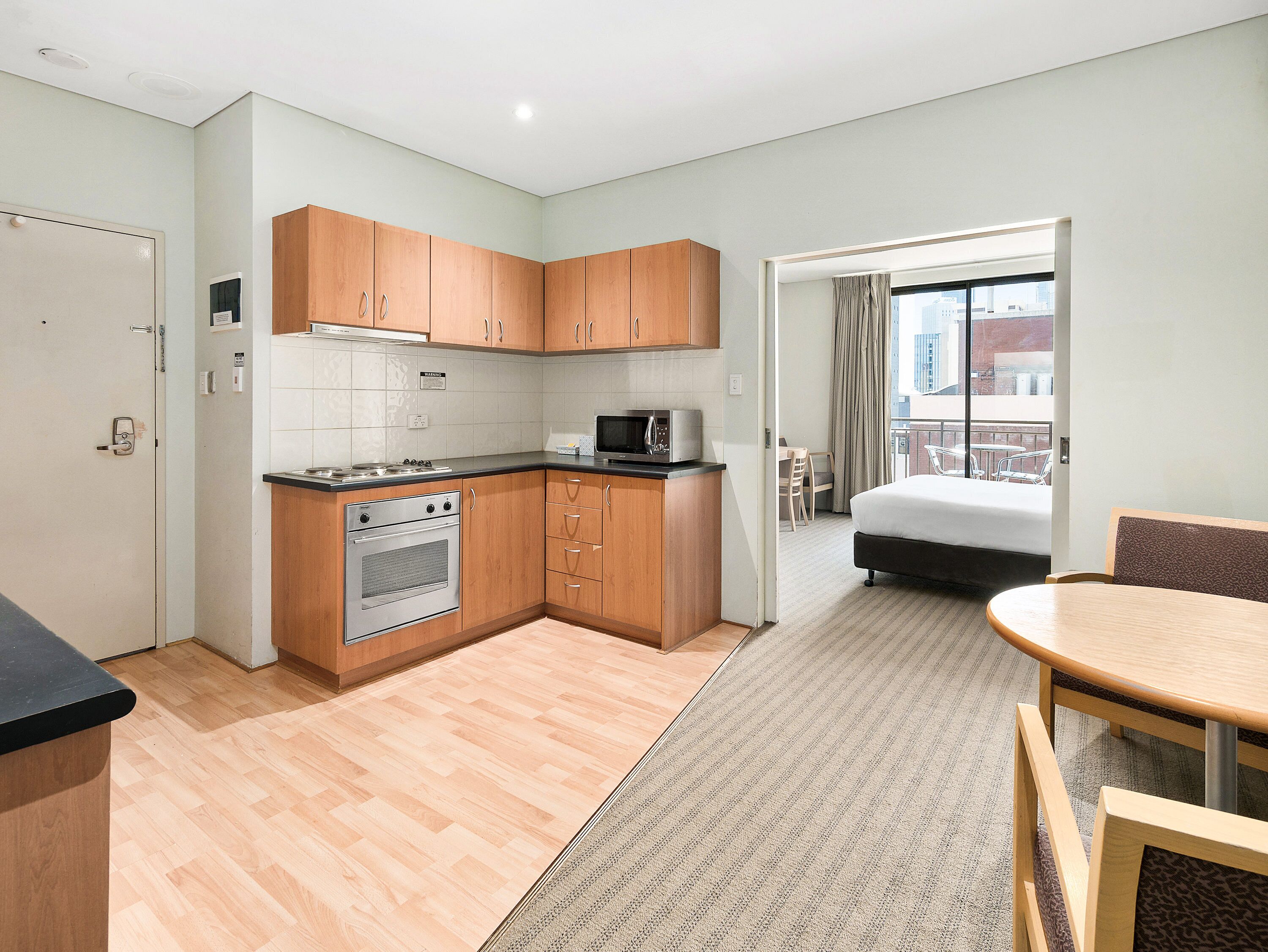 A compact hotel room with a kitchenette, a bed, a small dining area, and a view of the city through the window.