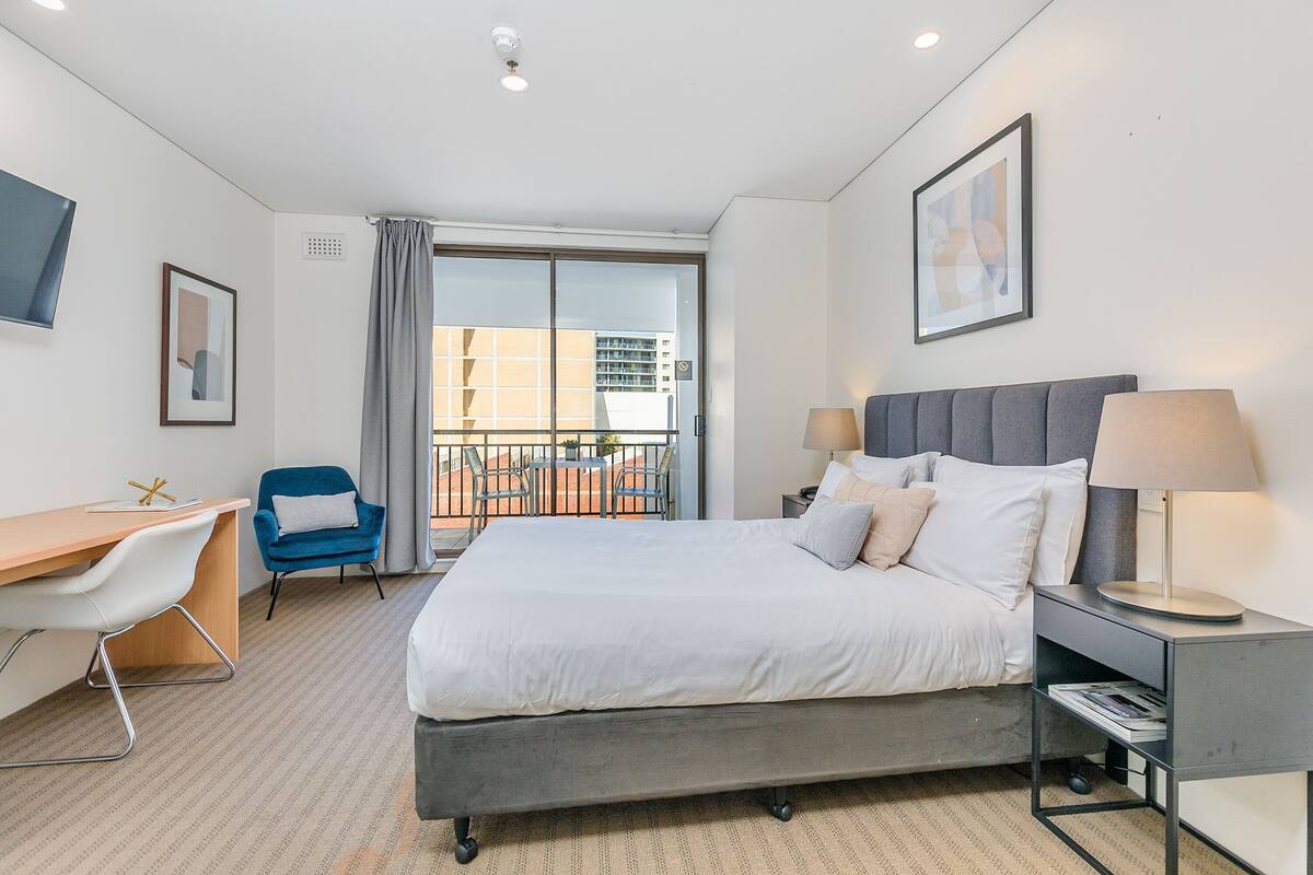 Habitación de hotel con cama, escritorio, silla, mesita de noche y vista al exterior.
