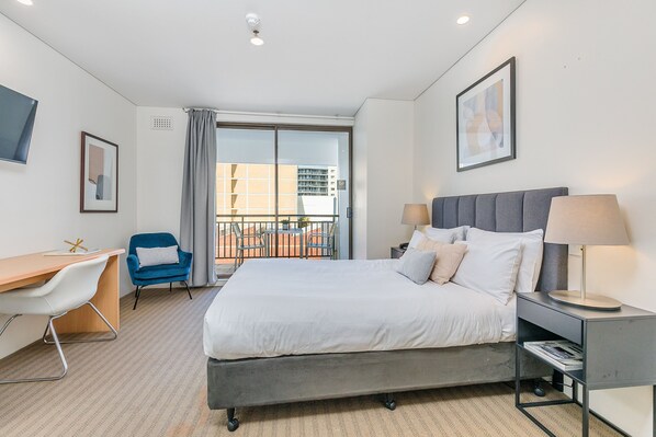 A hotel room with a bed, a desk, a chair, a nightstand, and a view of the outside.