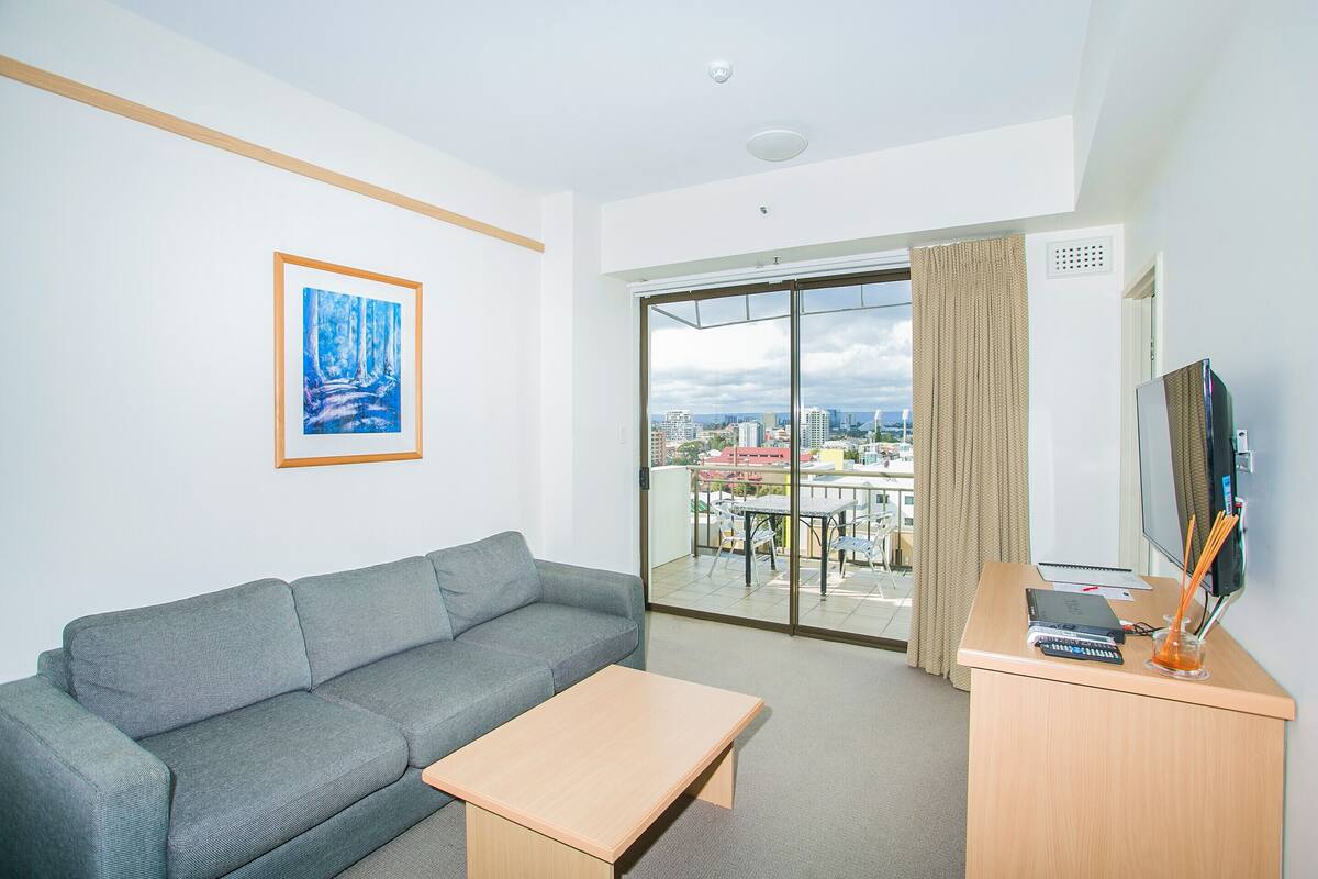 Una habitación de hotel moderna con un sofá gris, una mesa de centro de madera, un escritorio con una computadora, un televisor y un gran ventanal corredero que da a un balcón con vistas a la ciudad.