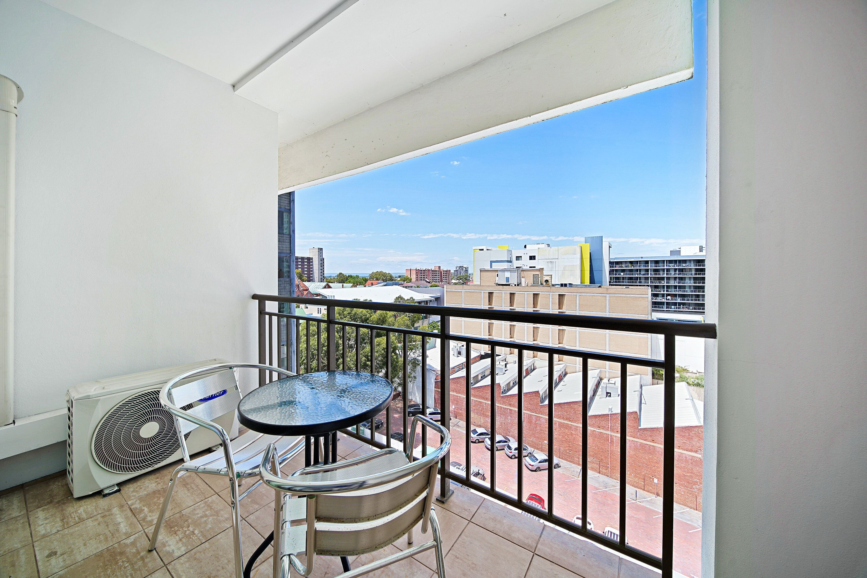 A balcony with a table and chairs, an air conditioning unit, and a view of buildings and a parking lot.