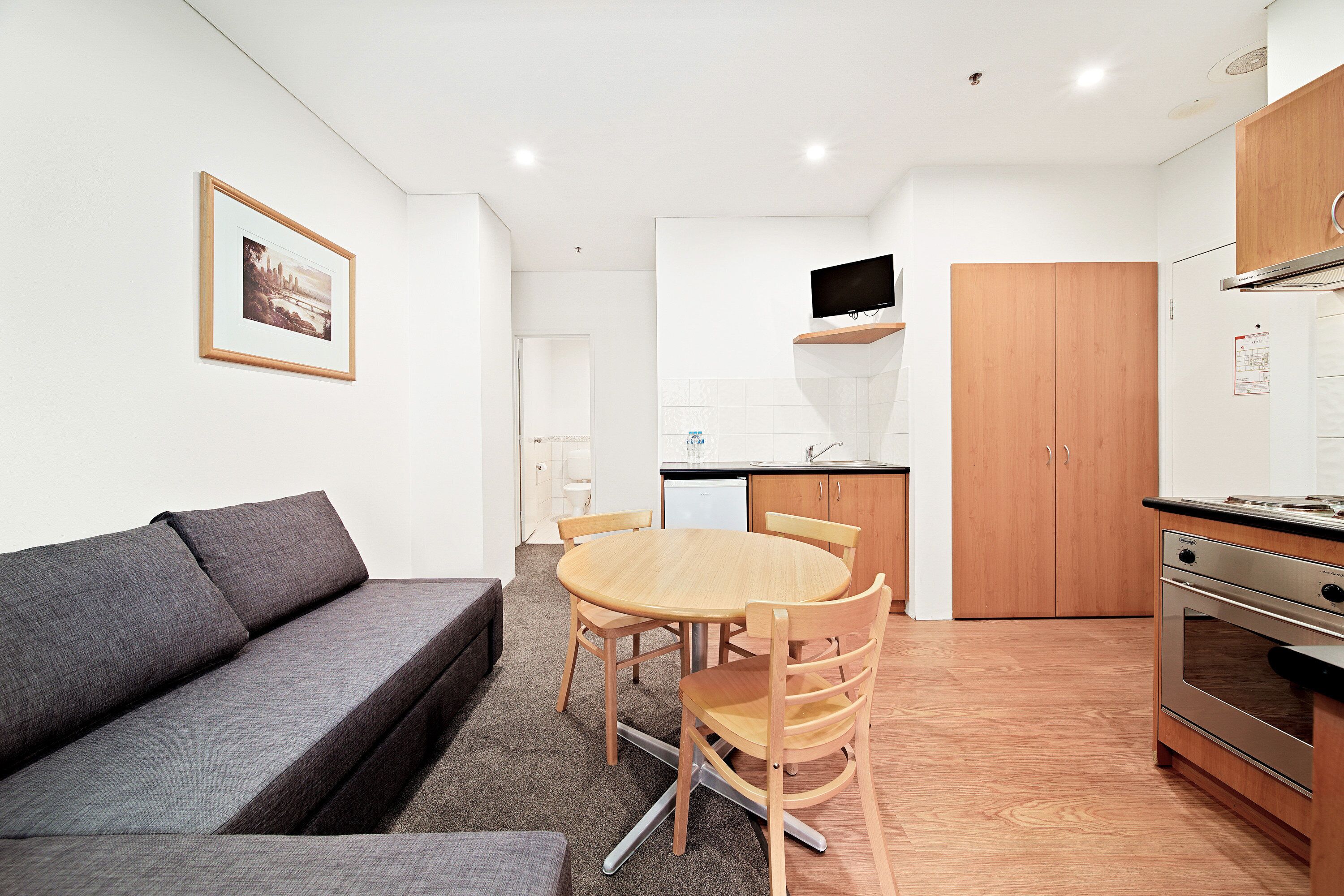 Ett kompakt bostadsutrymme med en soffa, ett matbord, stolar, ett köksområde med skåp och en inbyggd ugn samt en inramad tavla på väggen.