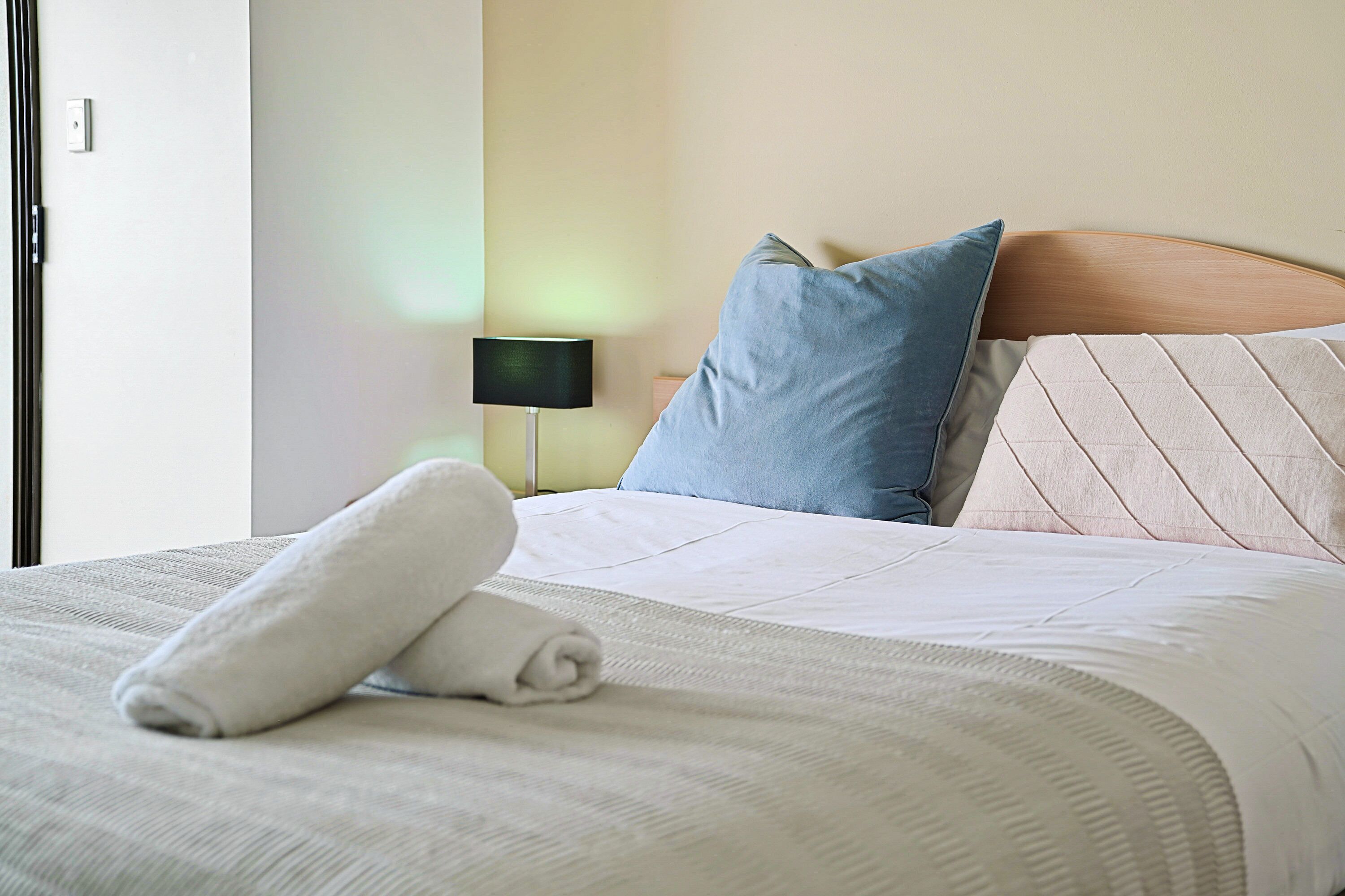 Un lit bien fait, avec une serviette blanche roulée, une tête de lit en bois et deux oreillers, un bleu et un beige.