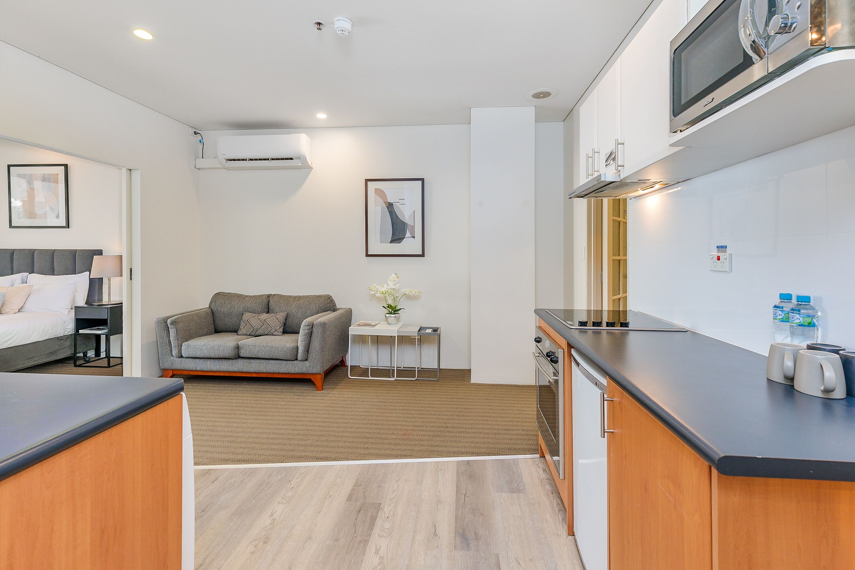 A compact hotel room with a kitchenette, a sofa, a small side table with a vase, and a framed picture on the wall.