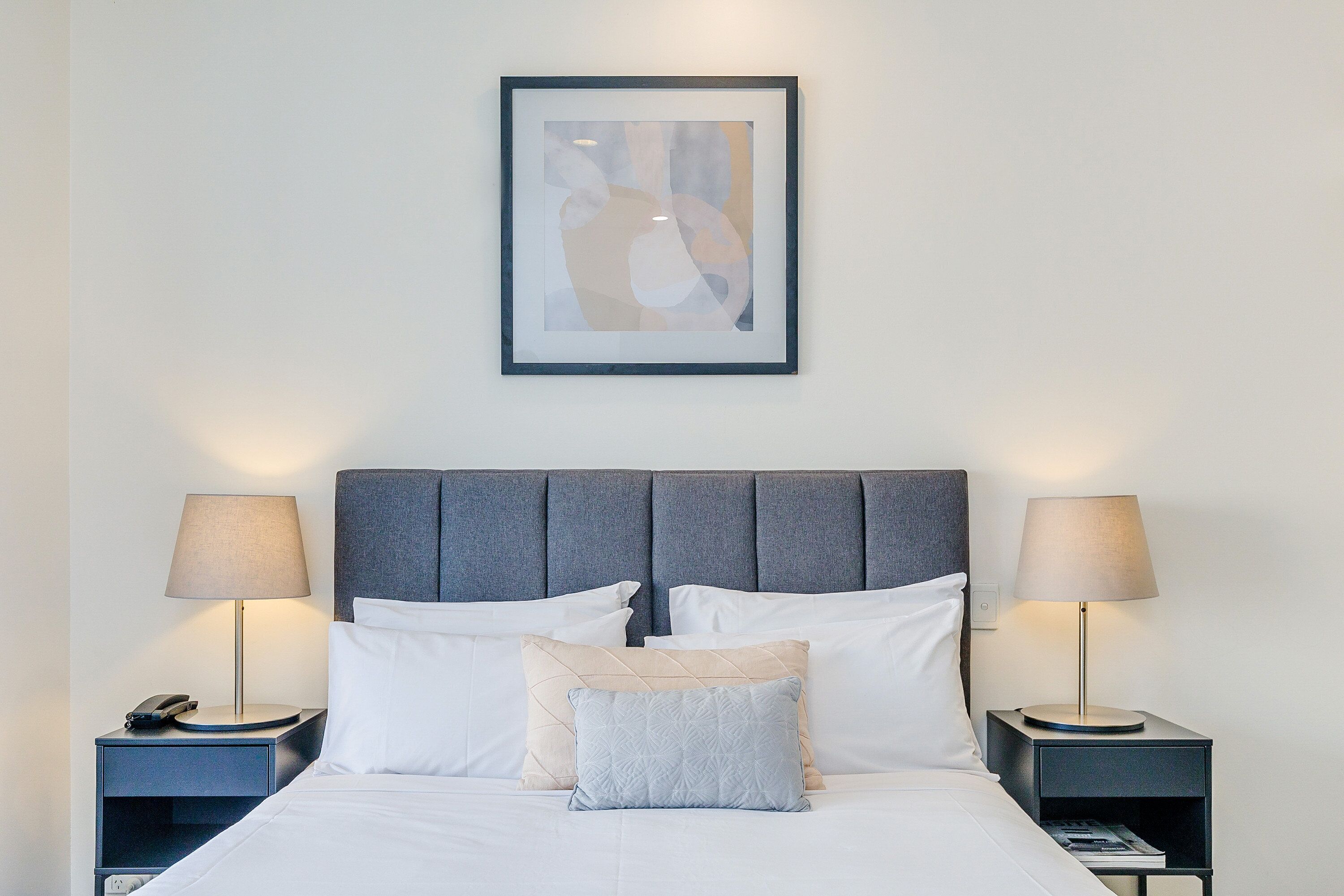 A neatly made bed with white linens, two bedside tables with lamps, and a framed abstract artwork above the bed.