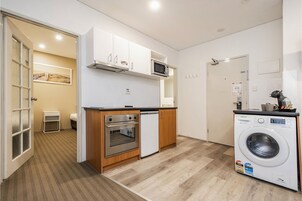 A compact kitchen with a microwave, oven, and dishwasher, a washing machine, and a small seating area with a chair and table.
