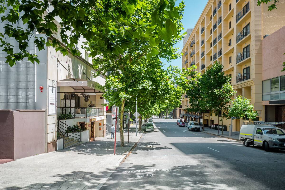 Una calle bordeada de árboles y edificios, incluyendo un edificio amarillo de varios pisos con balcones y un restaurante en la planta baja.