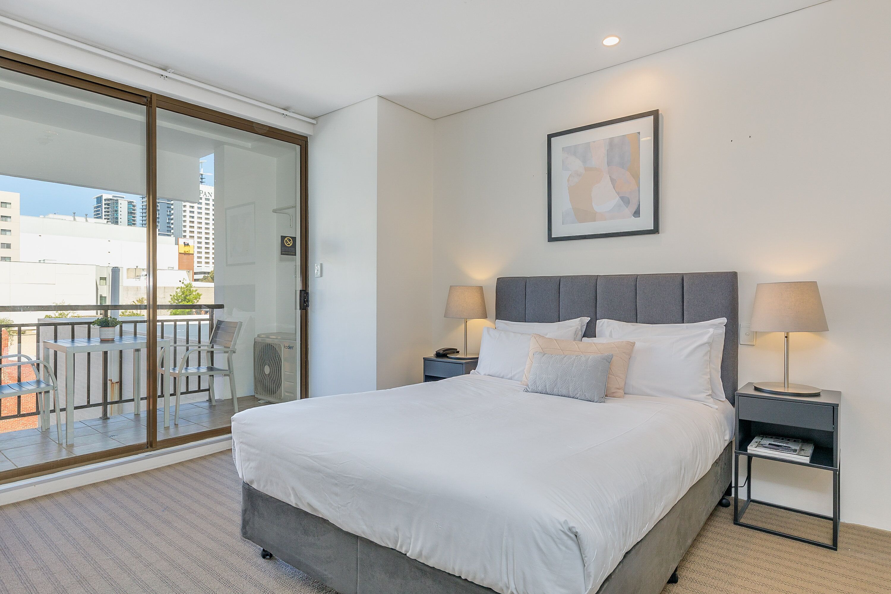 A modern hotel room with a large bed, two bedside tables with lamps, a sliding glass door leading to a balcony, and a framed abstract artwork on the wall.