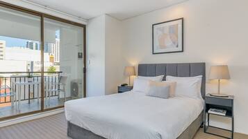 A modern hotel room with a large bed, two bedside tables with lamps, a sliding glass door leading to a balcony, and a framed abstract artwork on the wall.