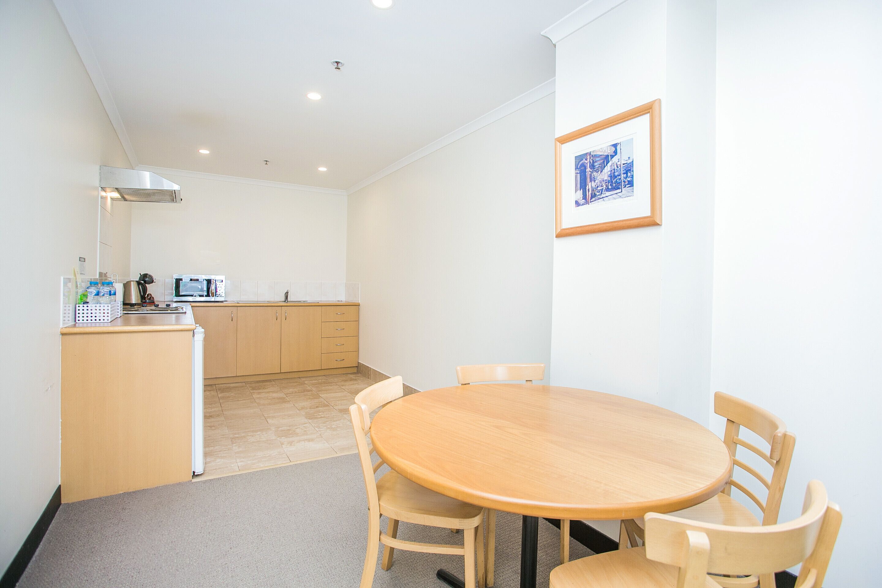 A small, clean room with a round wooden table and four chairs, a kitchenette with a microwave, and a framed picture on the wall.