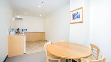 A small, clean room with a round wooden table and four chairs, a kitchenette with a microwave, and a framed picture on the wall.