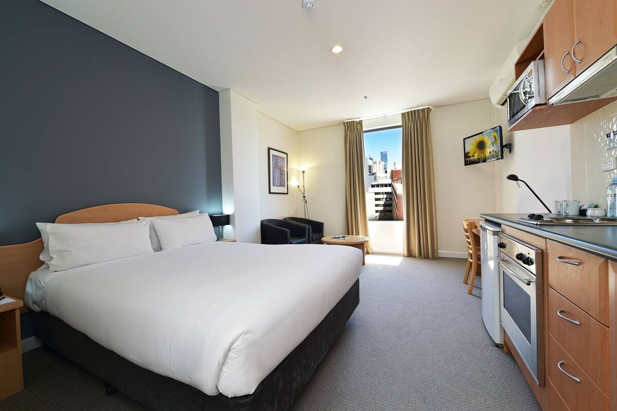 Habitación de hotel con una cama grande, kitchenette, una pequeña zona de estar y vistas a la ciudad desde la ventana.