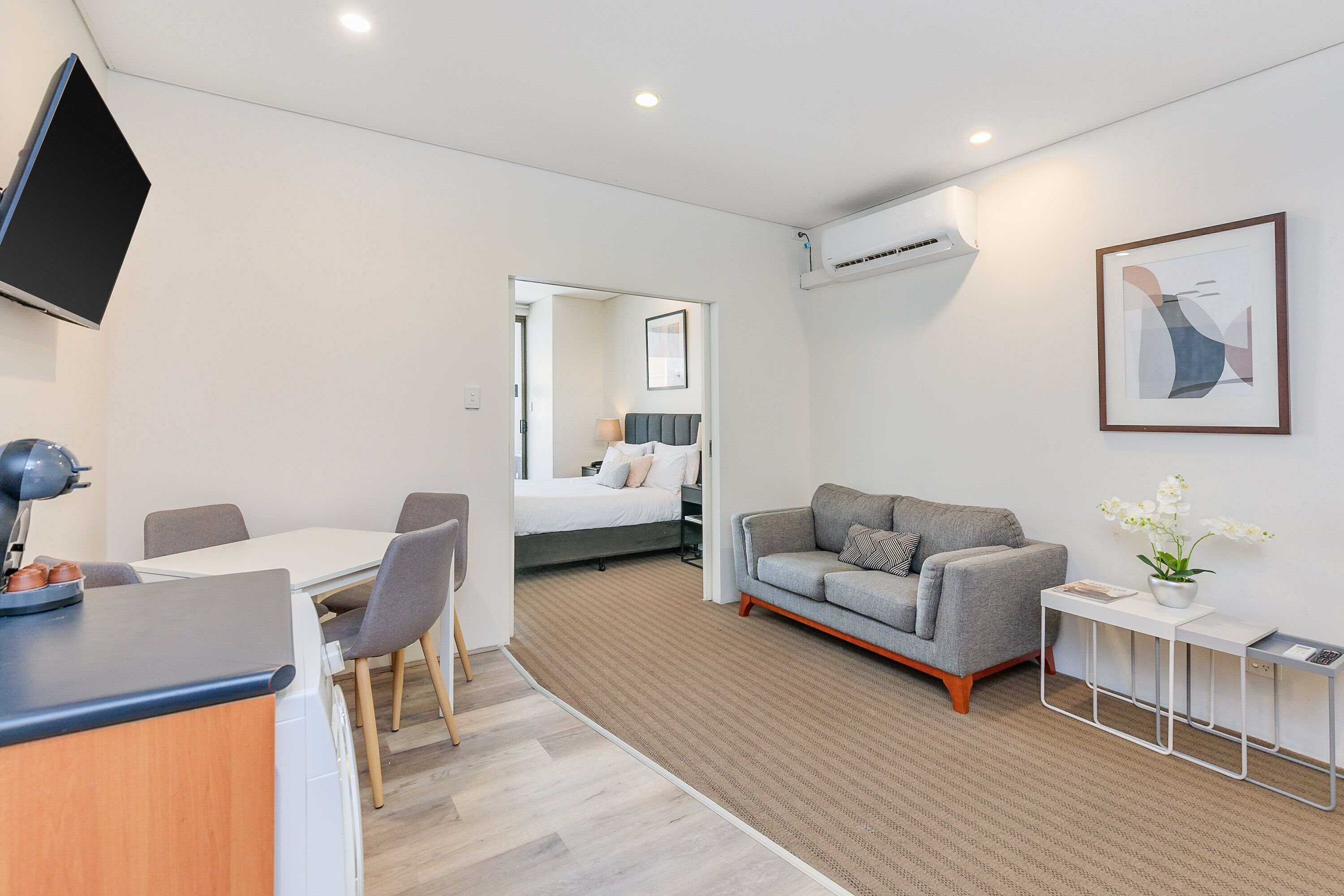 Une chambre d’hôtel moderne équipée d’un téléviseur à écran plat, d’un coin repas avec une table et des chaises, d’un canapé, d’une table de chevet avec une lampe et d’une œuvre d’art encadrée au mur.