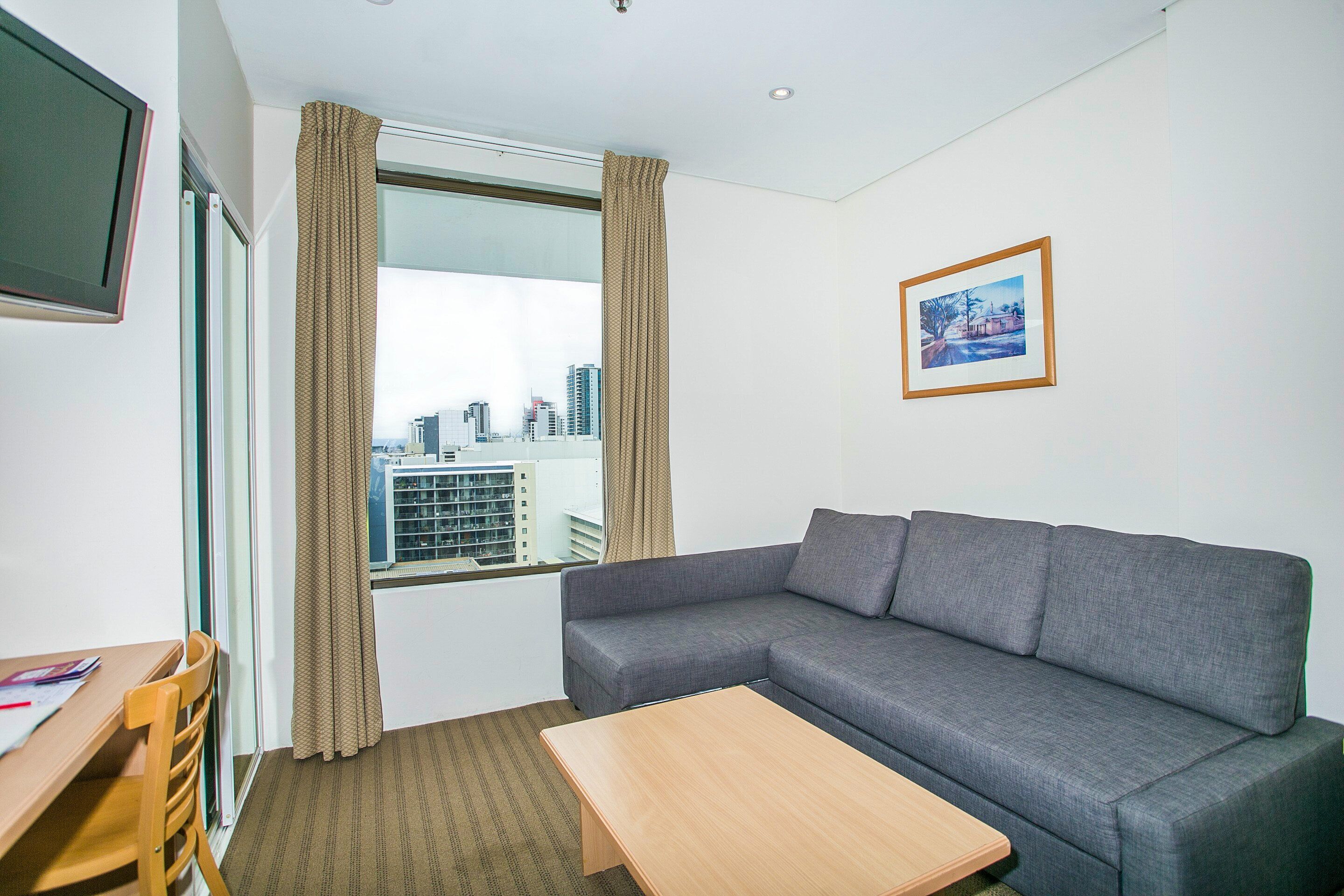 Una habitación de hotel moderna con un sofá gris, una mesa de madera, un televisor montado en la pared y una ventana con cortinas.
