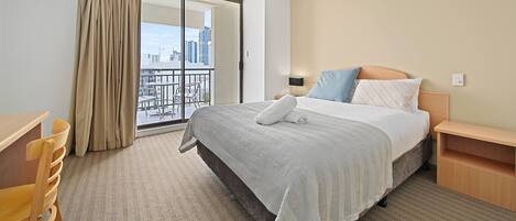 A hotel room with a bed, a desk, and a chair. There is a large window with a view of buildings.