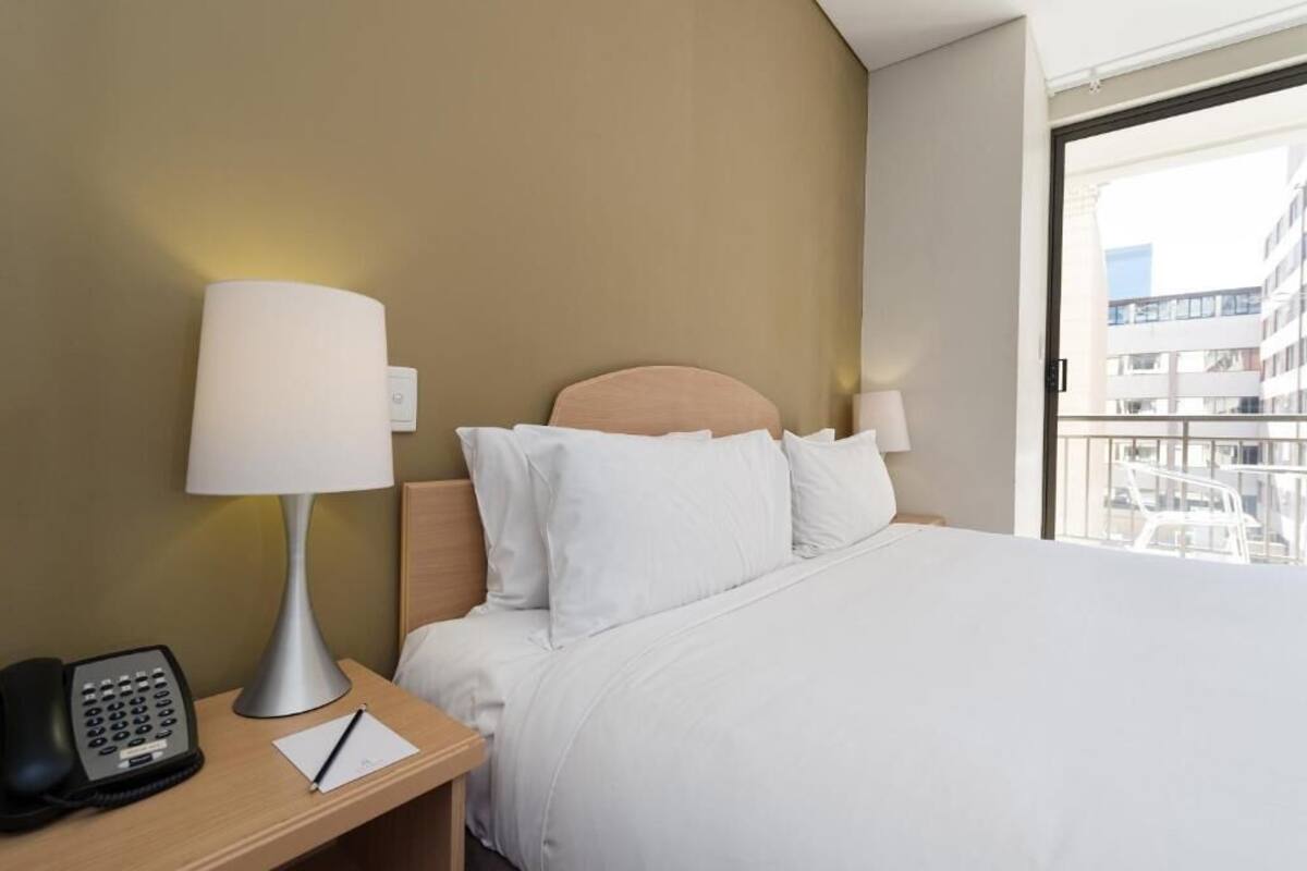 Habitación de hotel con cama, mesita de noche con lámpara, teléfono y ventana con vista a edificios.