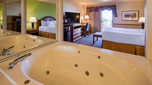 A hotel room with a large bathtub, a bed, a desk with a television, and a view of the outdoors. - Apple Tree Inn, SureStay Collection by Best Western (Petoskey)
