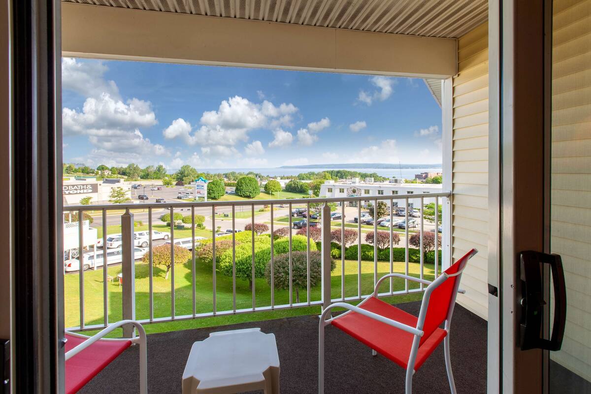 A balcony with two red chairs and a small table, overlooking a parking lot and greenery.