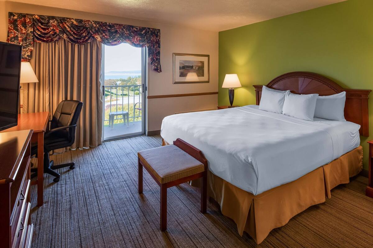 A hotel room with a large bed, two bedside tables with lamps, a desk with a chair, a small bench, and a view of the outdoors through a window with curtains.