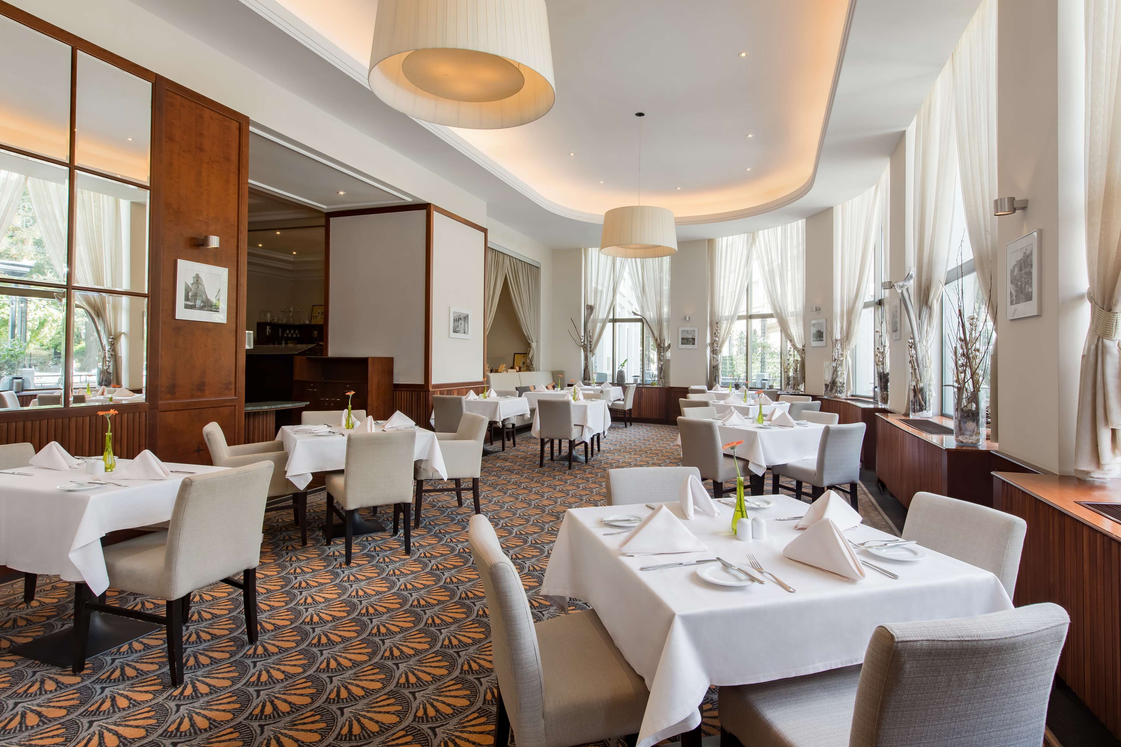 Ein geräumiges Restaurant mit mehreren Tischen und Stühlen, großen Fenstern mit Vorhängen sowie einem gestreiften Boden.