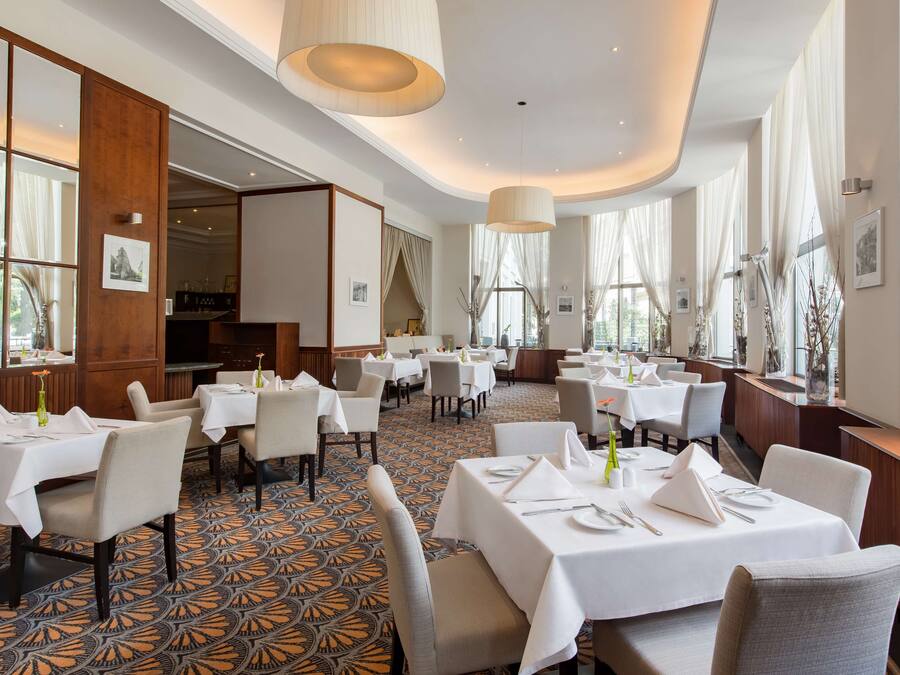 A spacious restaurant with multiple tables and chairs, large windows with curtains, and a patterned floor.